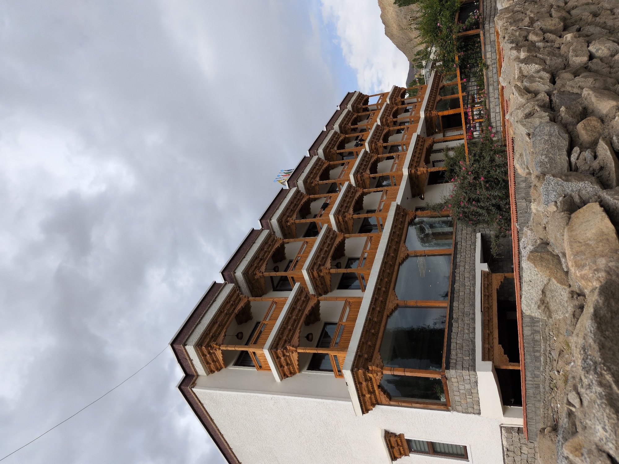 This is a low-angle, eye-level, full shot of a multi-story hotel in Leh, India, with a cloudy sky overhead. The hotel has a white stucco exterior with dark brown trim and intricately carved wooden balconies on multiple levels. The balconies have wooden railings and decorative wooden pillars supporting the overhangs. The first floor features large, reflective glass windows, likely for a lobby or restaurant. A low stone wall, constructed from rough-hewn rocks, runs along the foreground. Behind the hotel, a barren, mountainous landscape is visible, characteristic of Leh's high-altitude desert terrain. Some greenery, including flowering bushes, is present in front of the hotel. The overall scene depicts a tranquil, traditional architectural style set against a rugged natural backdrop.