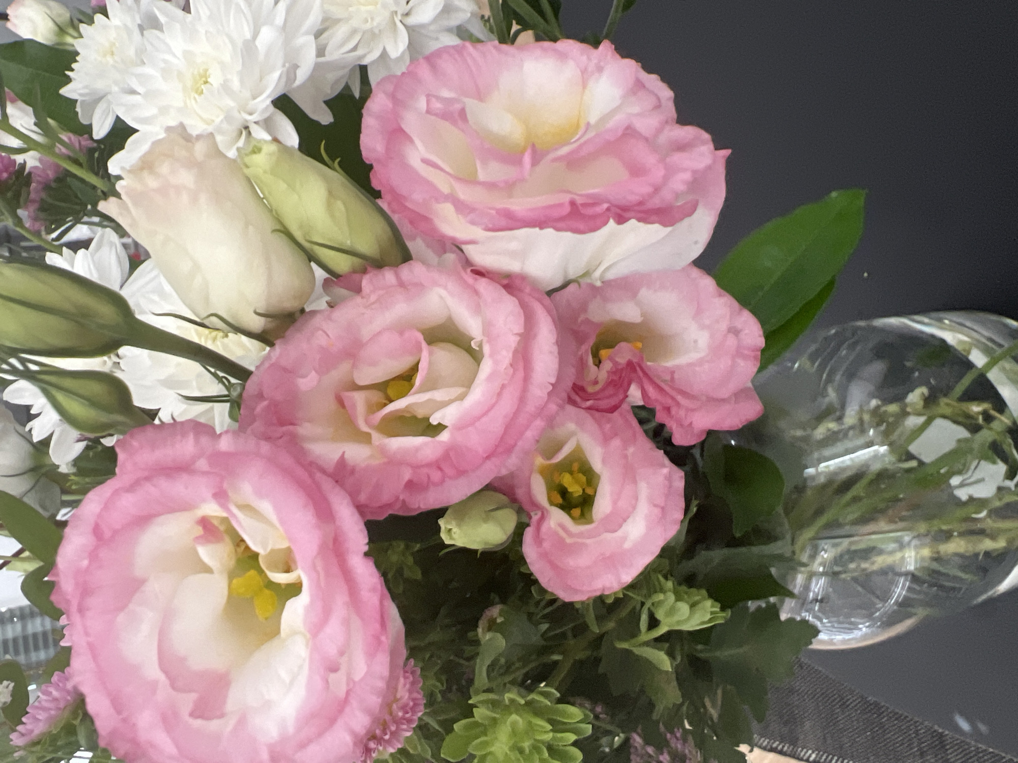 A close-up, top-down view of a bouquet of flowers. The primary subjects are pink and white lisianthus flowers and white chrysanthemums. The lisianthus flowers have delicate, ruffled petals that transition from white in the center to a soft pink at the edges, with yellow stamens visible in the open blooms. The chrysanthemums are a pure white, with many thin petals radiating from a central point. There are also several pale green buds and leaves interspersed among the flowers. To the right of the bouquet, a clear glass vase containing water and some greenery is partially visible. The background is a dark, out-of-focus surface, possibly a table or countertop. The lighting appears to be ambient, suggesting an indoor setting. There are no people visible, and no text is present in the image. The scene conveys a sense of natural beauty and freshness, characteristic of a flower arrangement.