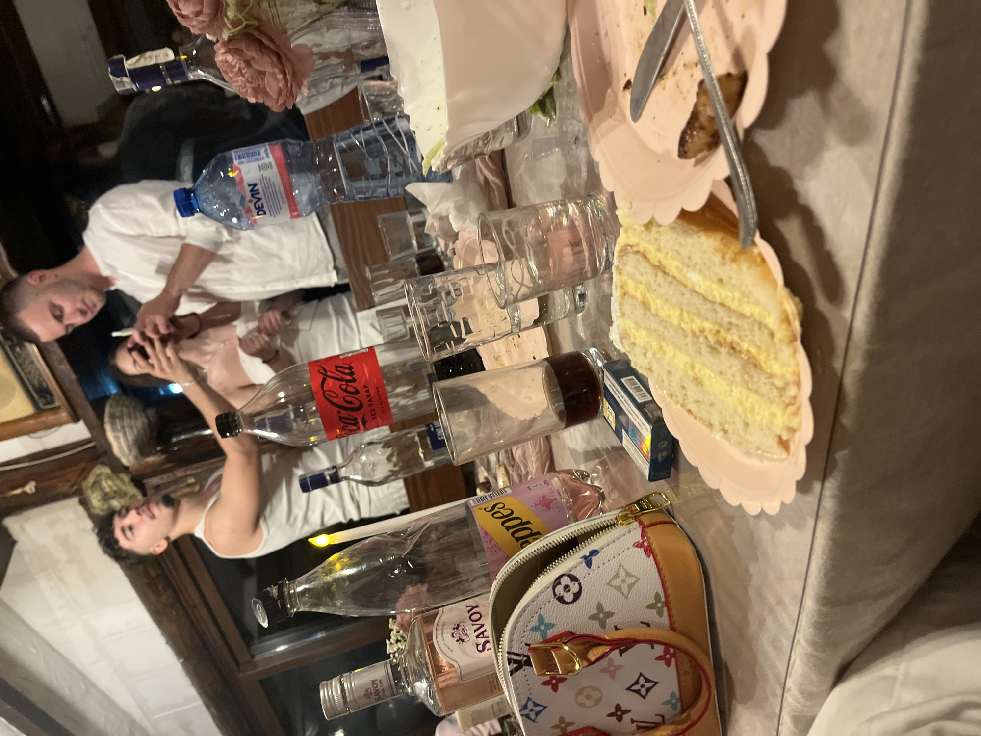 Here is a forensic description of the image:
**Scene:**
A tabletop cluttered with various items, seemingly after a gathering. Items include:
* Alcoholic beverages: Bottles of SAVOY (gin and rosé), FINLANDIA vodka, a clear bottle possibly containing spirits, and an opened Coca-Cola bottle (labelled "БЕЗ ЗАХАР БЕЗ КАЛОРИИ").
* Non-alcoholic beverages: A bottle of DEVÍN mineral water.
* Glassware: Several empty drinking glasses.
* Food: A slice of layered cake on a pink scalloped-edged plate with a knife. A plate with remaining crumbs of what looks like food.
* Personal item: A Louis Vuitton handbag (likely a small pochette or cosmetic case).
* A pack of cigarettes
**People:**
Two men and one woman are partially visible in the background.
**Objects:**
In addition to the objects noted above, a decorative floral arrangement is visible at the end of the table.
**Environment:**
The location appears to be indoors, possibly in a private home or a similar establishment. The city is presumable Presoka, Bulgaria based on the prompt. The lighting is relatively dim.
**Actions:**
The scene suggests a gathering where people have been consuming beverages and food.