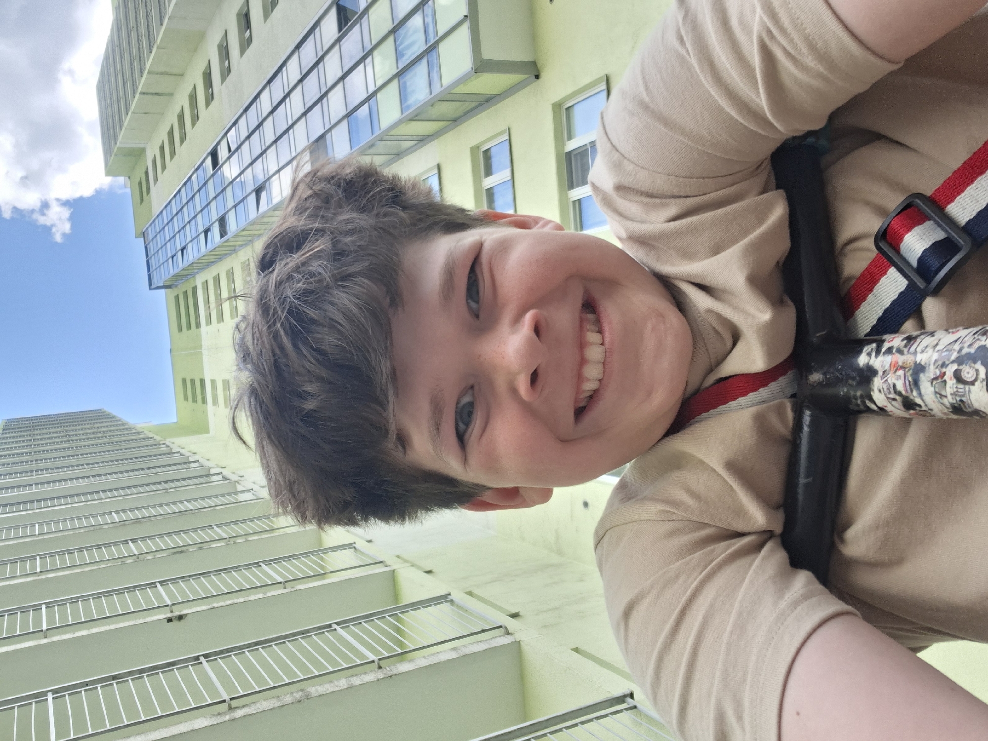 The image captures a young boy with short brown hair, fair skin, and blue eyes, smiling broadly directly at the camera. He is wearing a light beige short-sleeved top. A red, white, and blue striped strap with a black buckle is visible across his right side, possibly part of a bag or safety harness. The boy appears to be hanging upside down or leaning far over a black pole or bar, which has some indistinct patterns or stickers on its surface.
The photograph is taken from a low angle, creating an inverted perspective where the boy's face is prominent in the center-right foreground. The background features a tall, modern building with light green facades, characteristic of an urban setting in Saint Petersburg, Russia. The building extends vertically, showing multiple floors with balconies equipped with white railings on the left side of the frame. The upper section of the building on the right includes large, reflective windows that mirror the bright blue sky and scattered white clouds.
The clear blue sky with white clouds indicates it is daytime. The boy's wide smile and joyful expression suggest a playful or happy moment. No specific text is clearly legible, though faint patterns are visible on the black pole he interacts with.