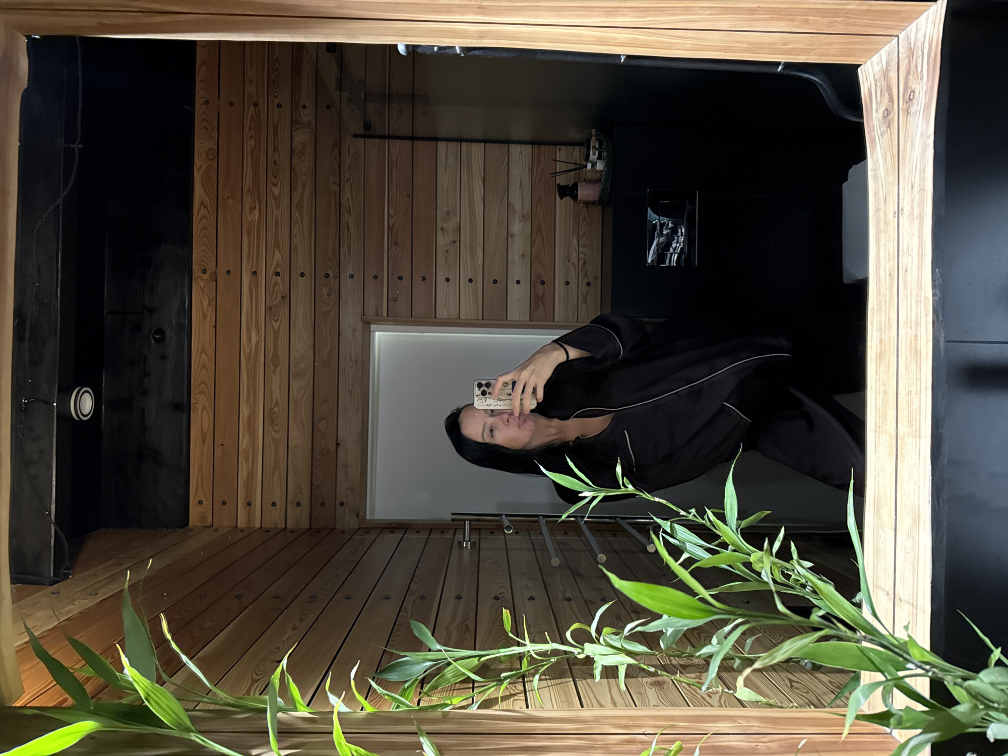 A woman with long dark hair takes a selfie in a bathroom with wood-paneled walls and a large mirror. She is wearing black pajamas and holding a smartphone. In the foreground, a green bamboo plant is visible. The bathroom appears to be part of a modern, possibly spa-like, setting. There is no indication of the city of Chaikovsky, Russia.