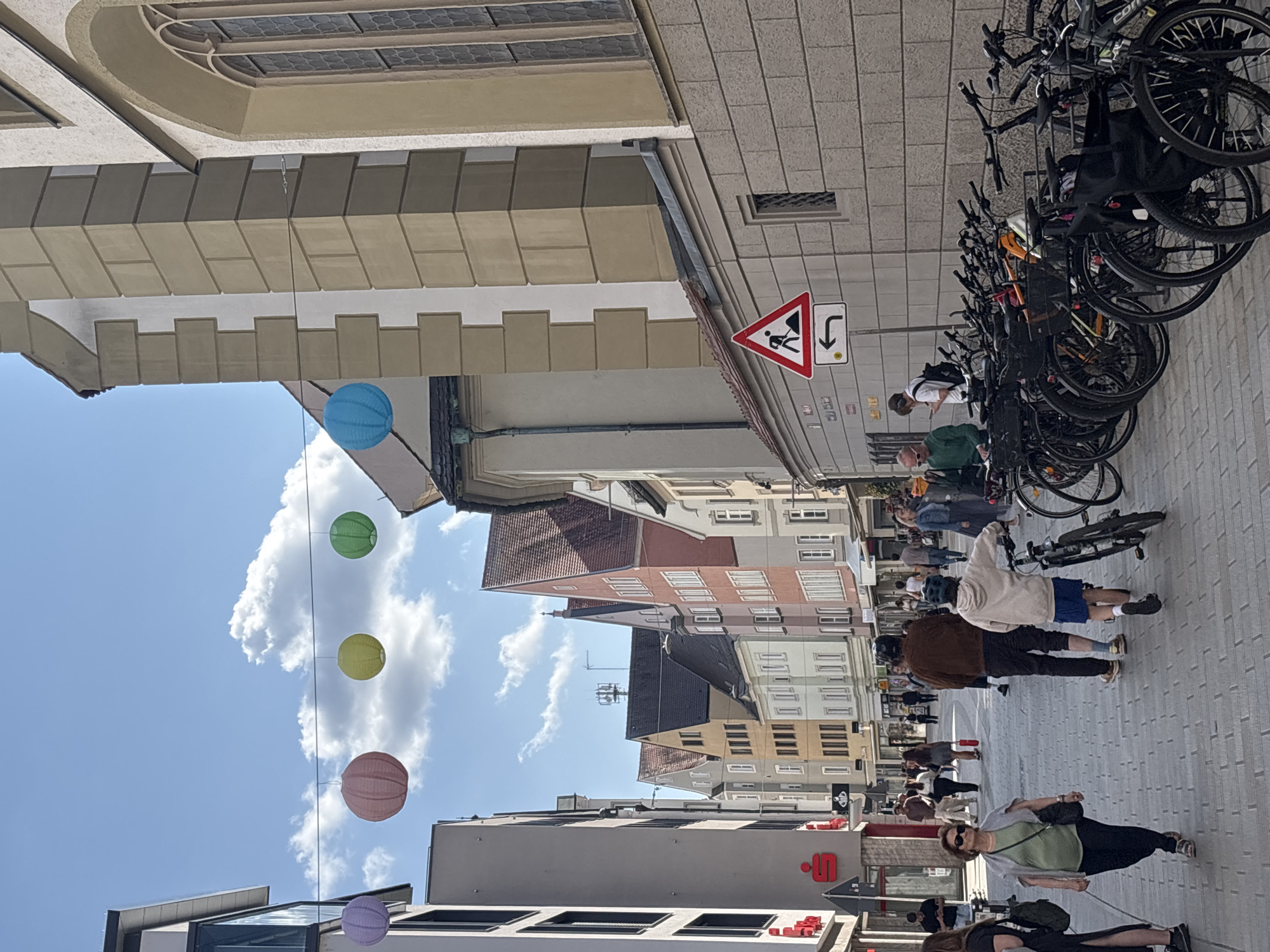 The image depicts a street scene in Augsburg, Germany. In the foreground, a paved pedestrian walkway is lined with parked bicycles on the right side. Several people are walking on the walkway. On the left, a modern building with large windows is visible, featuring a red logo that appears to be a "S" within a circle, characteristic of Sparkasse, a German savings bank. The background showcases a row of traditional European buildings with varied architectural styles and colorful facades. Overhead, a string of colorful paper lanterns hangs across the street, with a bright blue sky and scattered clouds above. A traffic sign indicating road work and a left turn is posted on the right side of the street. The overall scene suggests a bustling commercial or residential street in a European city.