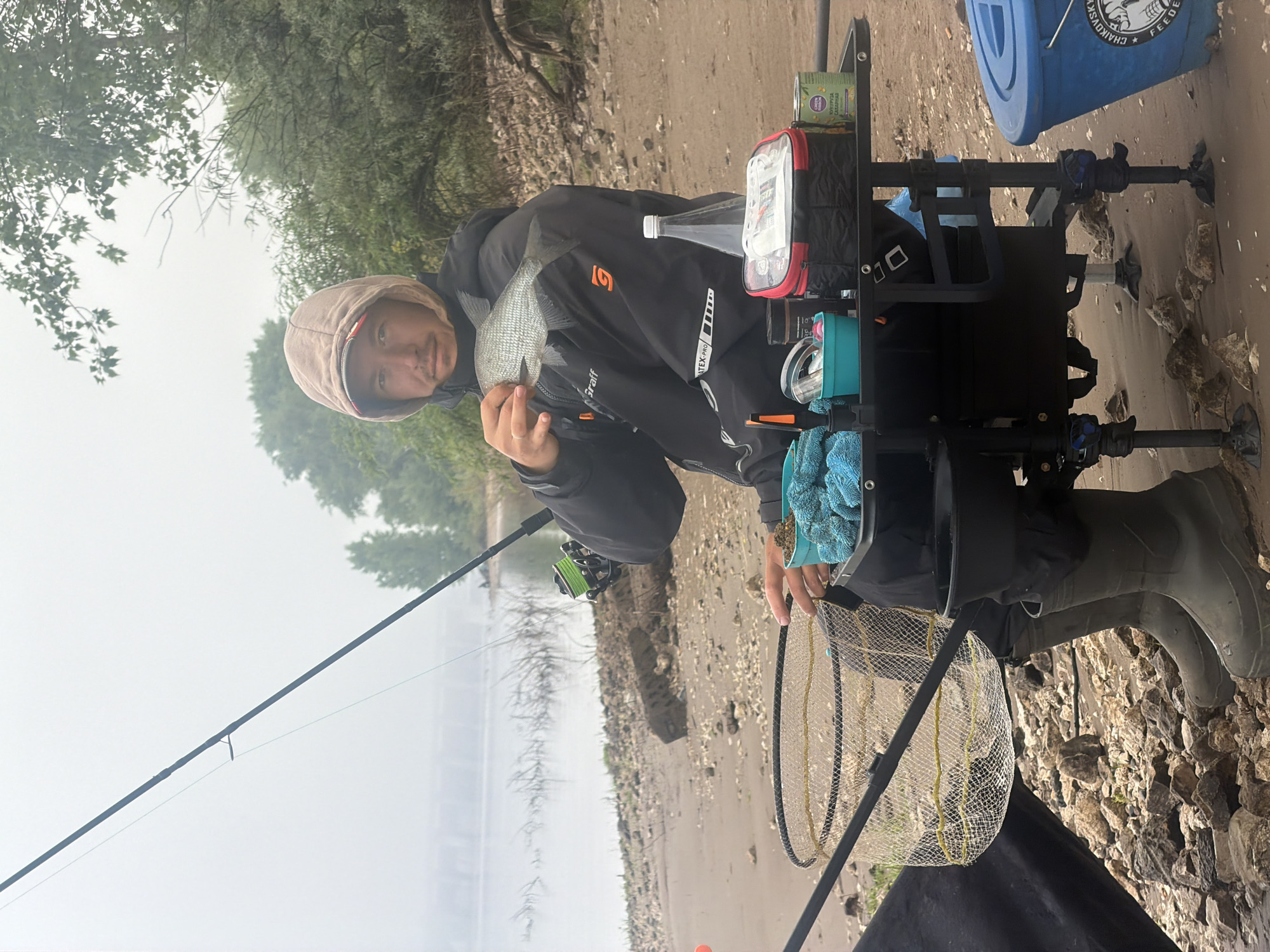 A vertical shot captures a male individual fishing on a rocky and sandy riverbank or lakeshore, likely in Chaikovsky, Russia, given the visible text. The weather appears overcast or hazy, with a wide body of water and treed distant banks contributing to a serene atmosphere.
The primary subject is a young man, wearing a dark hooded jacket with the hood up (light brown/beige) and dark pants, along with practical rubber boots. He is holding a freshly caught fish, possibly a bream or similar freshwater species, in his left hand, presenting it toward the camera with a subtle, content smile, indicating pride in his catch.
Arranged beside him is a black, multi-tiered modular fishing station or trolley. On its top shelf, a small red and black zip-up bag, a clear plastic water bottle, and a can are visible. The can's label shows Cyrillic text, including "Улов" (Ulov - Catch) and "Горошек зелёный" (Goroshek zelyonyy - Green peas), suggesting bait or food. A light blue container and a blue microfiber towel are also on the station. A fishing rod extends into the water from the left, fitted with a green reel. A circular landing net with a yellow mesh rests on the ground near his feet.
Further to the right and slightly behind, a blue bucket with a white lid is visible. Prominent text on the bucket reads "CHAIKOVSKY" and "FEEDER," accompanied by a stylized fish logo, linking the scene to the given location context. The man's jacket also displays "Graiff" and "LATEX PRO." The scene depicts a successful fishing outing during daytime, characterized by a quiet natural setting.