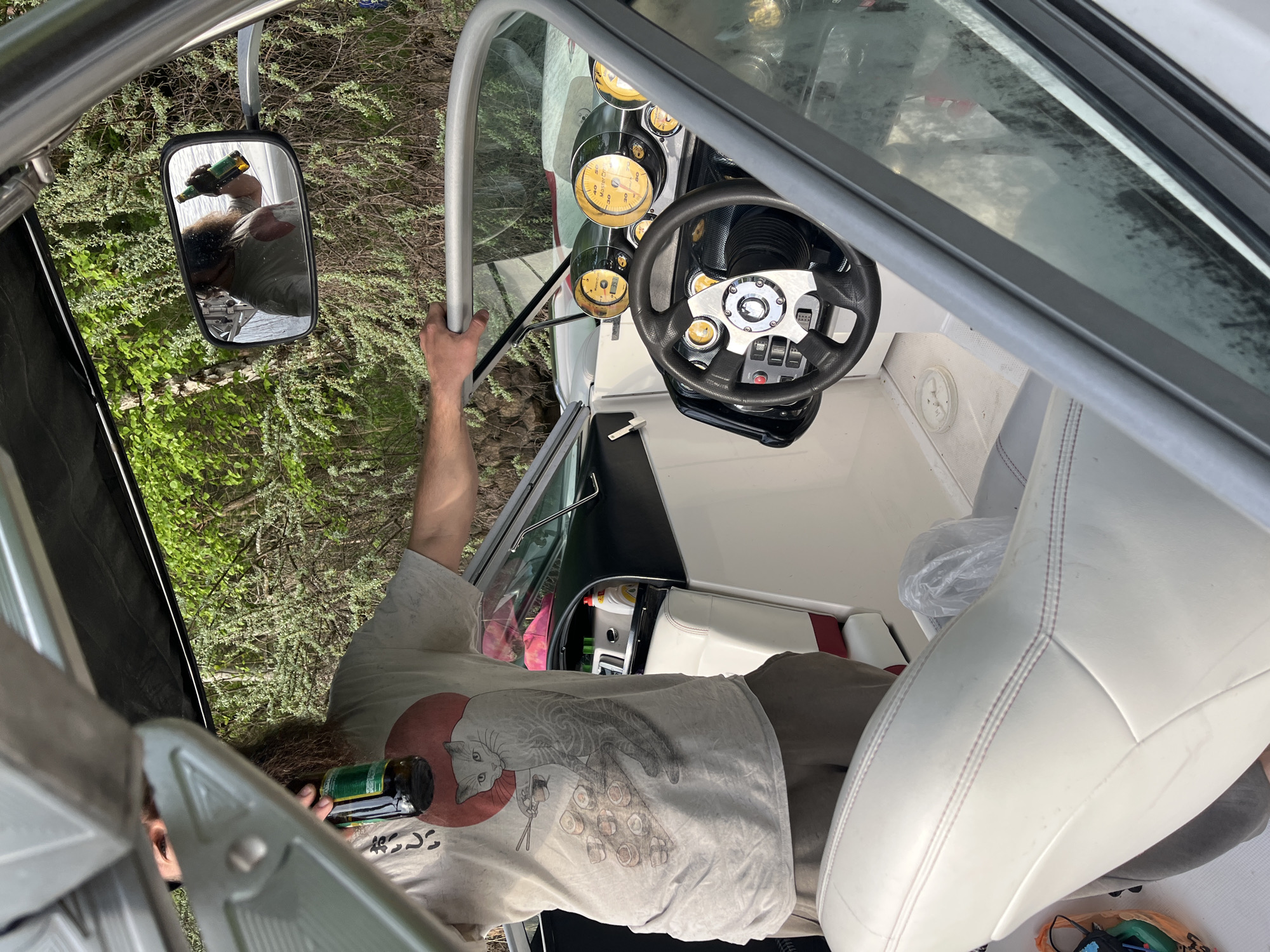 The image shows the interior of a boat, possibly a motorboat, with a fair-skinned individual holding a dark-colored bottle. The person is wearing a light-colored t-shirt with a graphic design. A mirror on the side of the boat reflects the person's face and the bottle, along with the surrounding natural environment. The boat's dashboard features various gauges and a steering wheel. The boat is white with interior upholstery. The environment outside the boat appears to be a forest or wooded area. It is not possible to confirm the location as Пятница, Russia from the image alone.