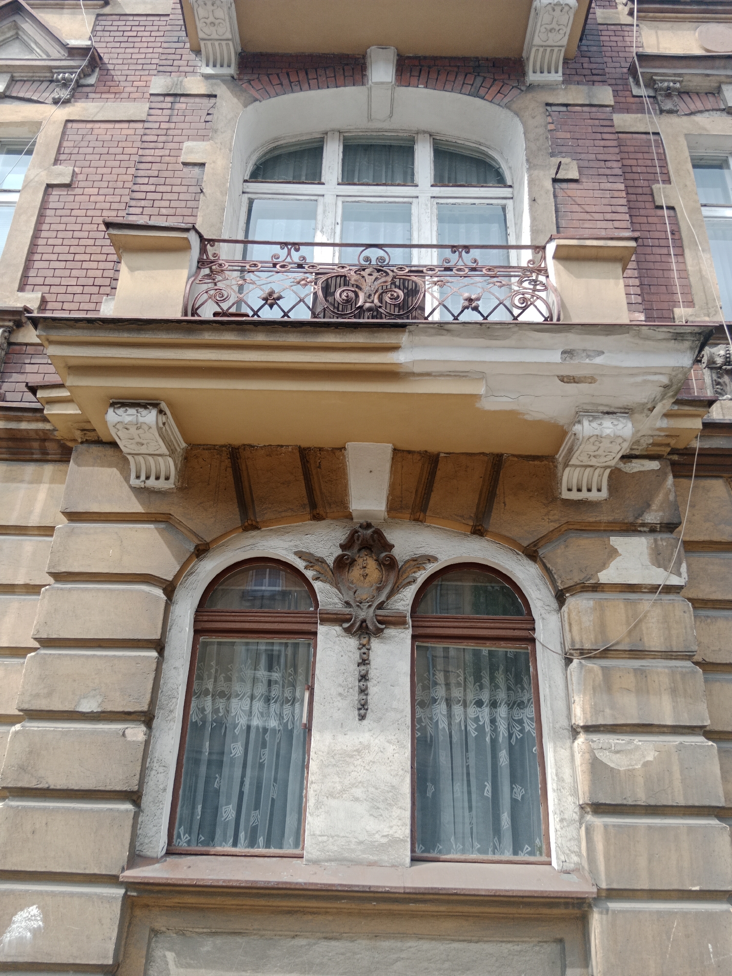 An ornate, arched balcony with decorative iron railings extends from a brick facade of a building. Below this balcony, two arched windows are visible, each covered with sheer white curtains. The building's exterior displays a mix of brickwork and stone elements, showing signs of age and weathering. The image appears to be taken from a low angle, looking up at the architecture. The building is located in Bydgoszcz, Poland.