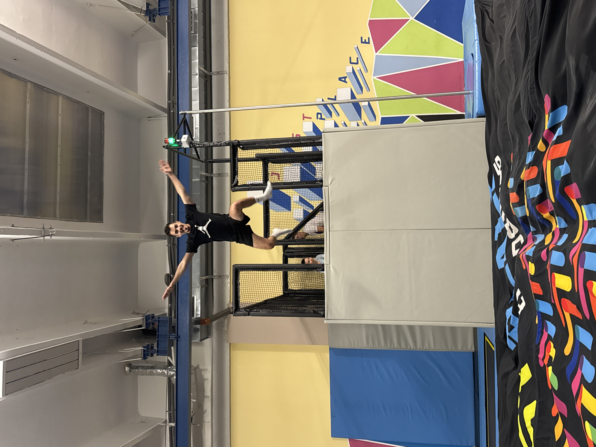 An action shot captures a man mid-air within an indoor trampoline park or adventure facility. The man, wearing a black t-shirt with a white Jordan logo, black shorts, and white socks, is suspended horizontally, smiling broadly with his arms and legs outstretched in an acrobatic pose. He appears to be in the process of jumping or swinging.
The setting includes a vibrant yellow wall with stylized text spelling "TRACER PLACE" and colorful geometric patterns in blue, pink, and green. Below the man, a black netted enclosure or soft play structure is visible, with another person's head peeking out. Light gray padded walls and boxes surround the activity area, and a blue trampoline surface is also present. To the right, a black fabric banner with bright, abstract colorful shapes and partial letters like "JUMP" adds to the lively decor. The ceiling and upper walls are white, featuring industrial vents and structural elements. The scene conveys an energetic and fun recreational activity taking place indoors, likely in Sofia, Bulgaria.