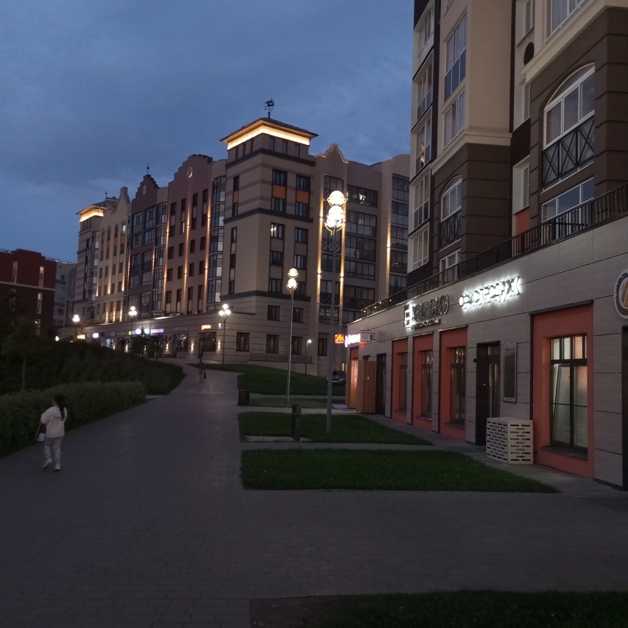 The image depicts a modern residential and commercial complex at dusk in Izhevsk, Russia. The architecture features multi-story buildings with a mix of beige and dark brown facades, accented by numerous lit windows and external lighting strips. Several shops with illuminated signage are visible on the ground floor. A paved walkway lined with green lawns and trees leads through the complex. One person, a woman dressed in light-colored clothing, is walking away from the camera on the walkway. The sky is a deep blue, indicating the transition from day to night.