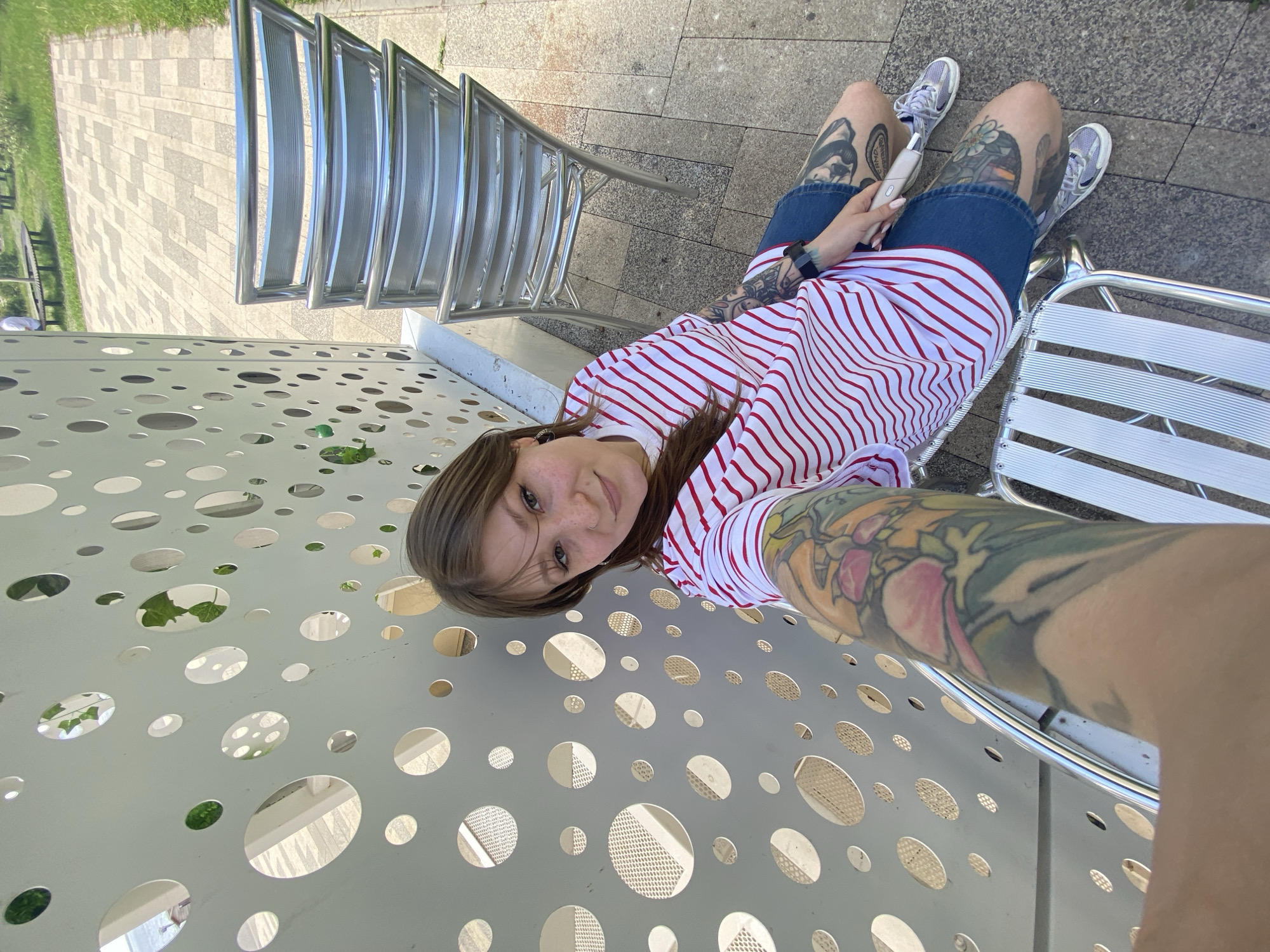 A medium shot depicts a young woman with fair skin, light brown hair, and freckles, lying on her side on a perforated metal surface. She is wearing a red and white striped shirt and denim shorts. Her legs are visible, revealing numerous tattoos. She is also wearing grey sneakers. Her left hand holds a white object. Her right arm, heavily tattooed, extends towards the camera.
The location appears to be an outdoor urban setting, evidenced by metal chairs, a tiled walkway, and greenery in the background. While the request is to identify the location in Saratov, Russia, there is not enough detail in this image to ascertain the exact location.