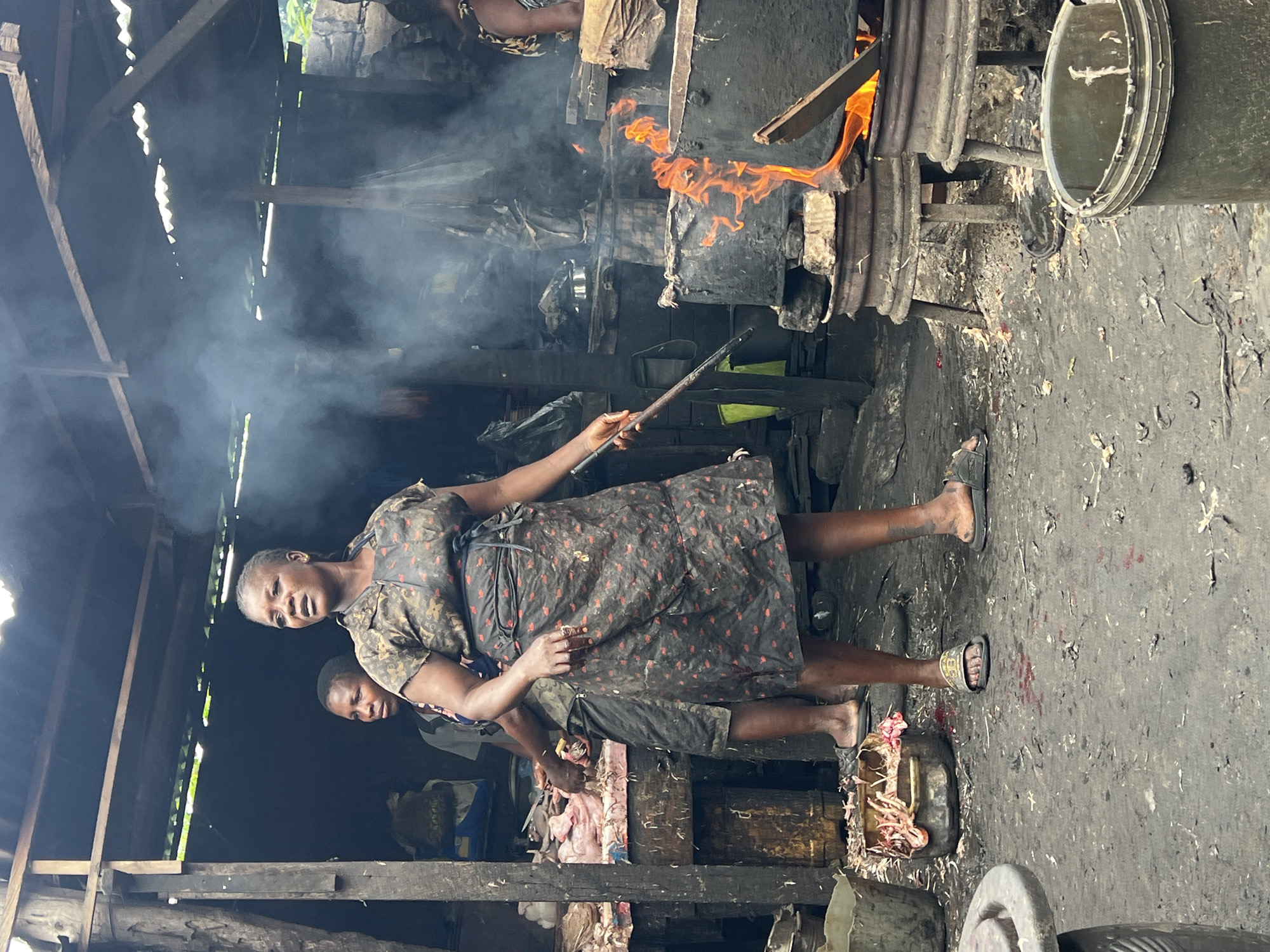 This media file depicts an outdoor scene in Nkpor, Nigeria, likely a marketplace or a workshop. The setting appears to be rustic and somewhat industrial, with a wooden structure overhead and a dirt floor covered in debris. A prominent feature is a large fire burning in a metal structure, producing a significant amount of smoke that fills the upper portion of the frame.
The primary subject is a woman standing in the foreground, wearing a dark, patterned dress and flip-flops. She is holding a long stick, possibly for tending the fire or stirring a pot. Her expression suggests engagement with the activity. Behind her, another person is partially visible, also appearing to be involved in the work.
The activity taking place appears to be cooking or some form of processing involving the fire. The presence of what looks like raw meat in a container suggests food preparation. The overall atmosphere is one of bustling activity, typical of a busy work environment.
Notable details include the dense smoke, indicating the intensity of the fire, and the worn appearance of the surroundings, hinting at a long-standing operation. The lighting suggests it could be daytime, but the dense smoke obscures direct sunlight. There is no visible text in the image.