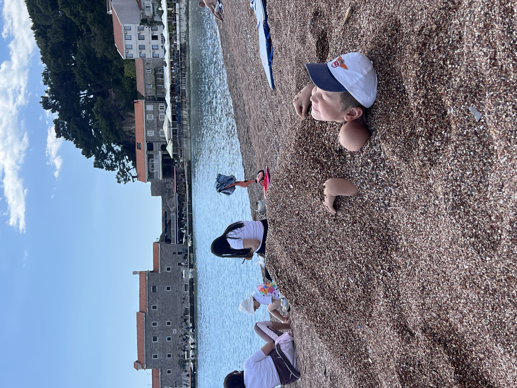 A child is buried in pebbles on a beach, wearing a baseball cap, with only their head, hands, and upper chest exposed. Other people, including children and adults, are on the same beach near the water's edge. The Petrovac centar, Montenegro coastline and associated buildings are in the background.