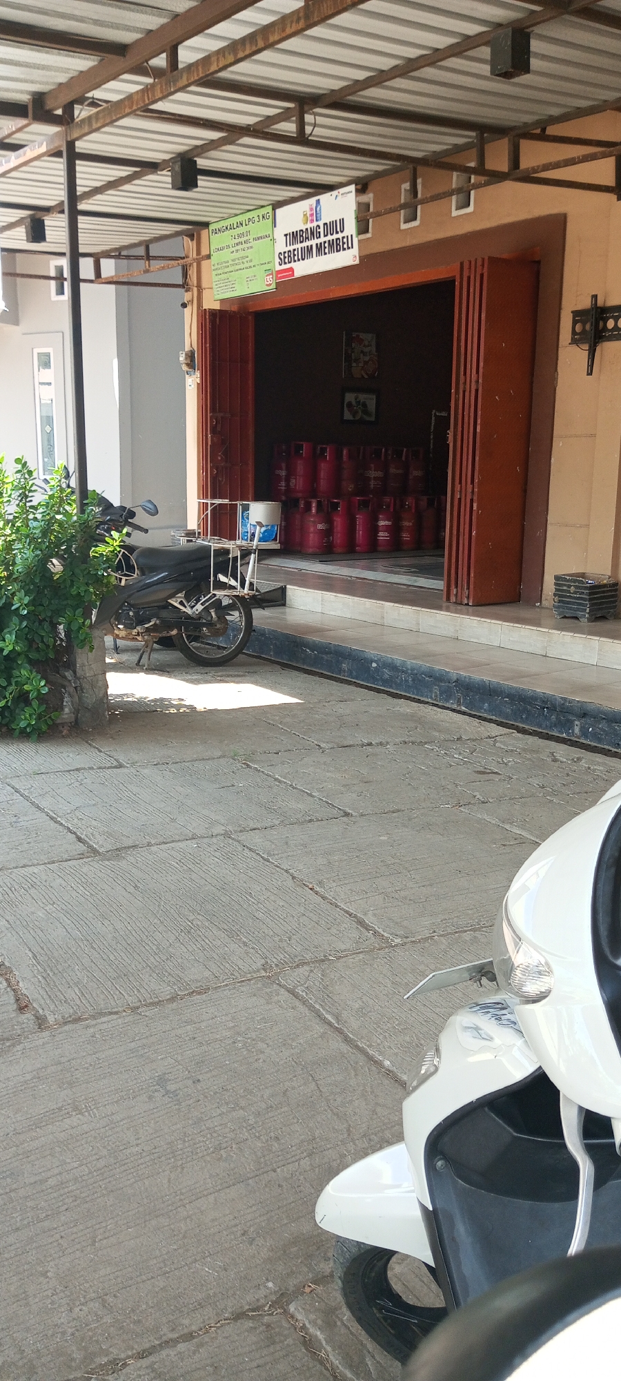 The image shows a front view of a gas station for LPG 3 kg in the city of Lempa, Indonesia. The building has a corrugated metal roof supported by a metal frame. On the facade, there are two signs. One sign, predominantly green, displays information about "PANGKALAN LPG 3 KG" and lists a location as "JL. LEMPA NO.1, PAMMANA" with a phone number. The second sign, in white and red, reads "TIMBANG DULU SEBELUM MEMBELI" which translates to "Weigh first before buying".
Inside the open building, numerous red LPG cylinders are neatly stacked. In front of the building, a black motorcycle with a white metal rack attached to its rear is parked on a concrete pavement. The pavement is laid with square concrete slabs that have a textured, grooved surface. To the right of the building's entrance, a white scooter is partially visible in the foreground. A large green bush is located to the left of the motorcycle. The scene appears to be under bright daylight, casting shadows on the ground.