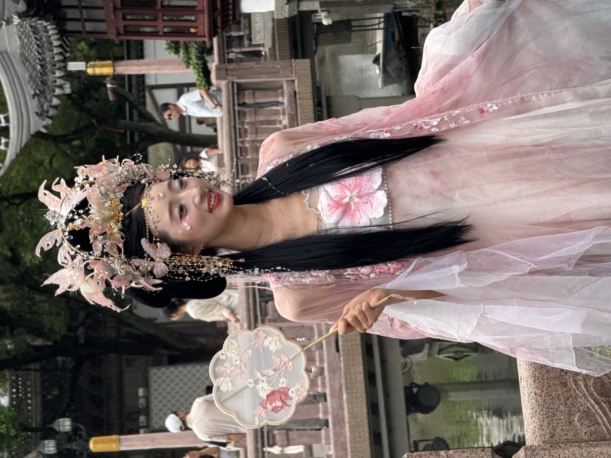 A young woman, dressed in a pink and white traditional Chinese outfit and elaborate headwear, stands on a stone bridge, holding a fan. She is smiling and looking to her right. The background suggests a park or garden setting, with water, trees, and traditional architecture. People can be seen in the distance, some walking, others observing. The overall scene appears to be a tourist attraction or cultural event, possibly in Shanghai, China.