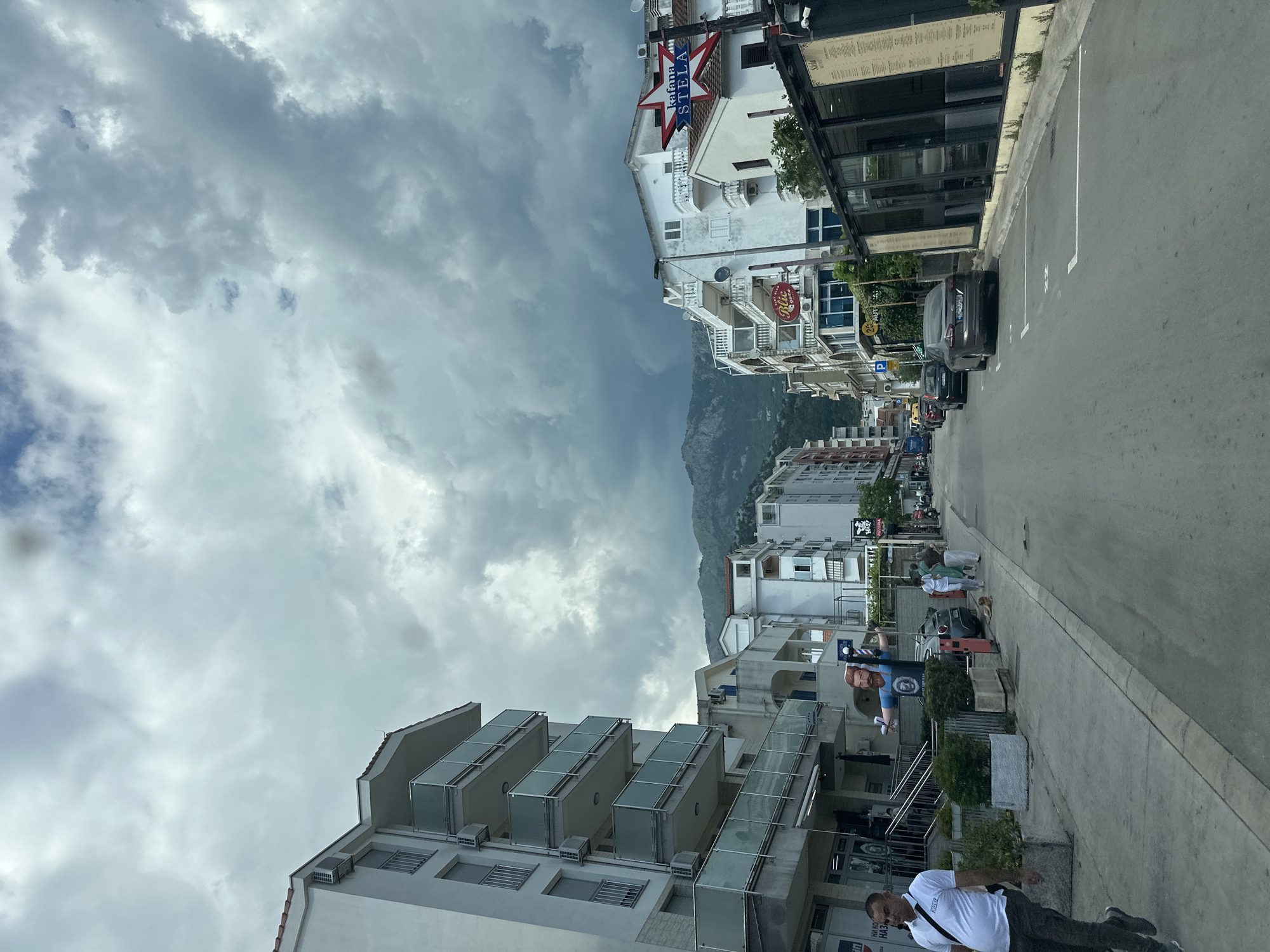 The image depicts a street in Budva, Montenegro, under a cloudy sky. Buildings line both sides of the street. The buildings are mainly white and multi-storied, with balconies. A mountain is visible in the distance between the buildings. On the right side of the street, there is a row of parked cars. People are walking along the street. A man in a white shirt and dark pants is walking towards the camera on the right side of the street. A barber shop sign is visible, suggesting the presence of a barber shop nearby.