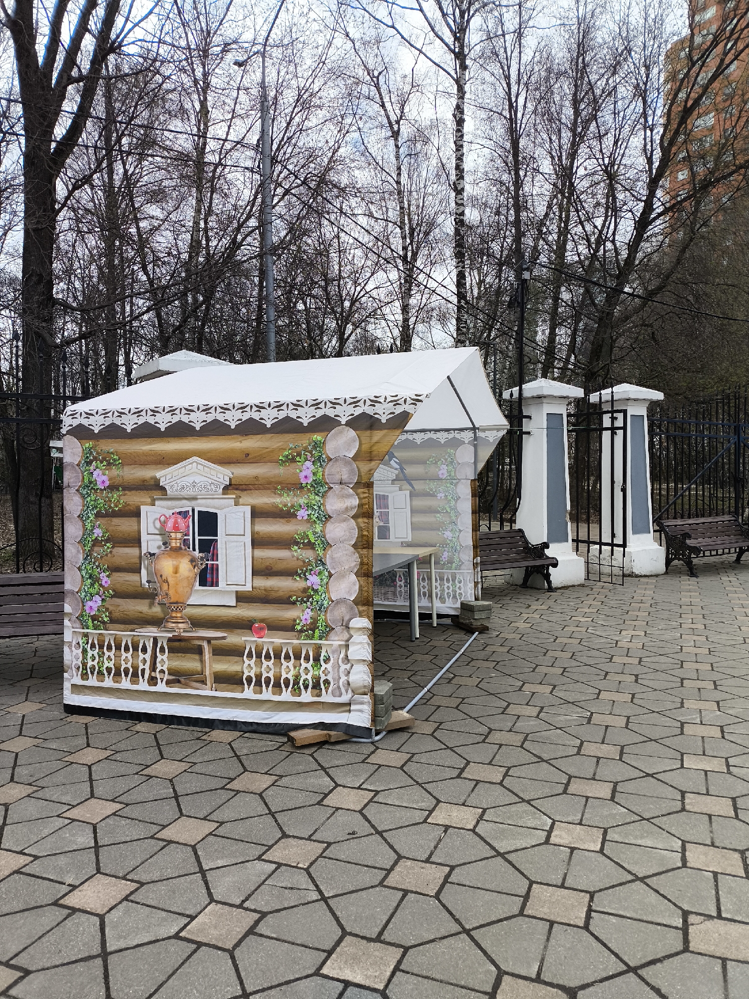 In Khimki, Russia, a temporary structure resembling a traditional Russian log cabin stands on a patterned paved surface. The structure is printed with a design of a log cabin facade, complete with a window, a samovar, and floral decorations. A white canopy covers the top of the structure. Behind the structure is a black metal fence with white and gray pillars, two park benches, and a backdrop of bare trees and a multi-story building. The sky is overcast.