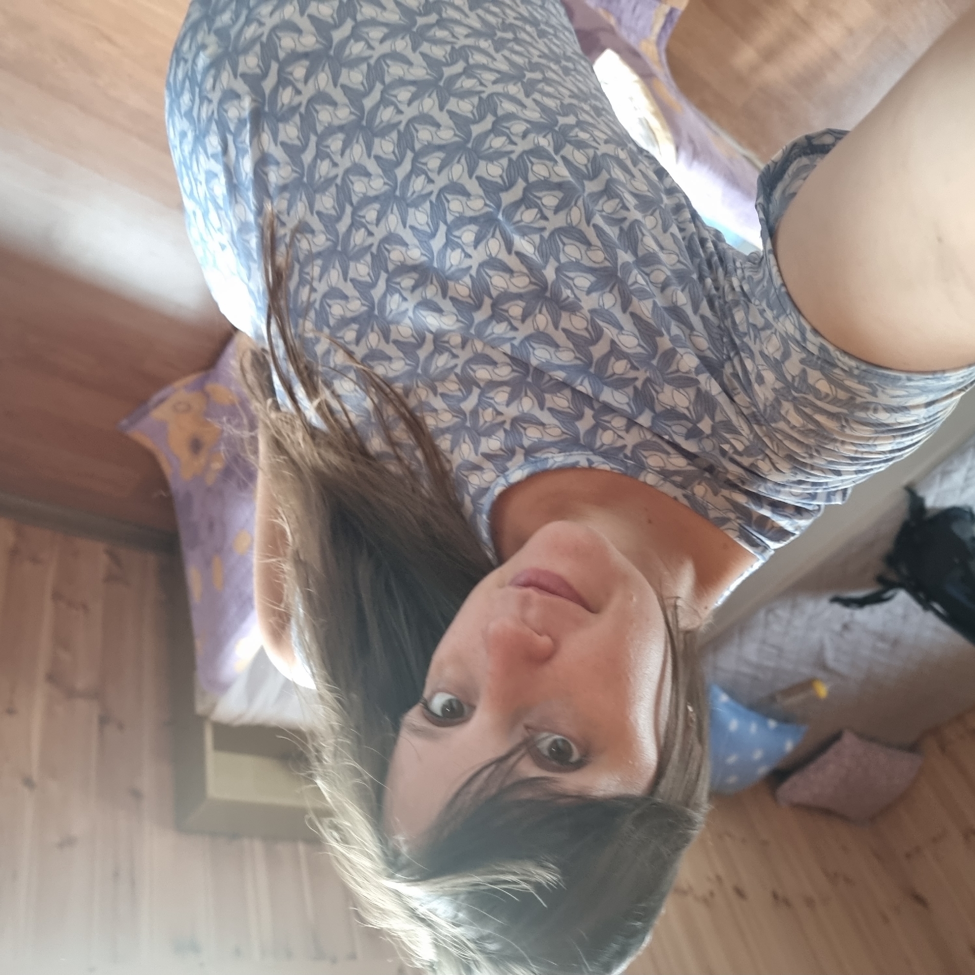 An overhead, close-up shot shows a young woman with long brown hair, wearing a light blue t-shirt with a repeating white and blue floral pattern. She is in a room with light wood paneled walls and a wooden floor. To the left of the woman, a bed is visible with grey and light blue patterned bedding, and two pillows. One pillow is light blue with white dots, and the other is light pink. A black bag is on the left side of the bed. To the right of the bed is a purple blanket with a yellow floral pattern. The scene appears to be in a bedroom.