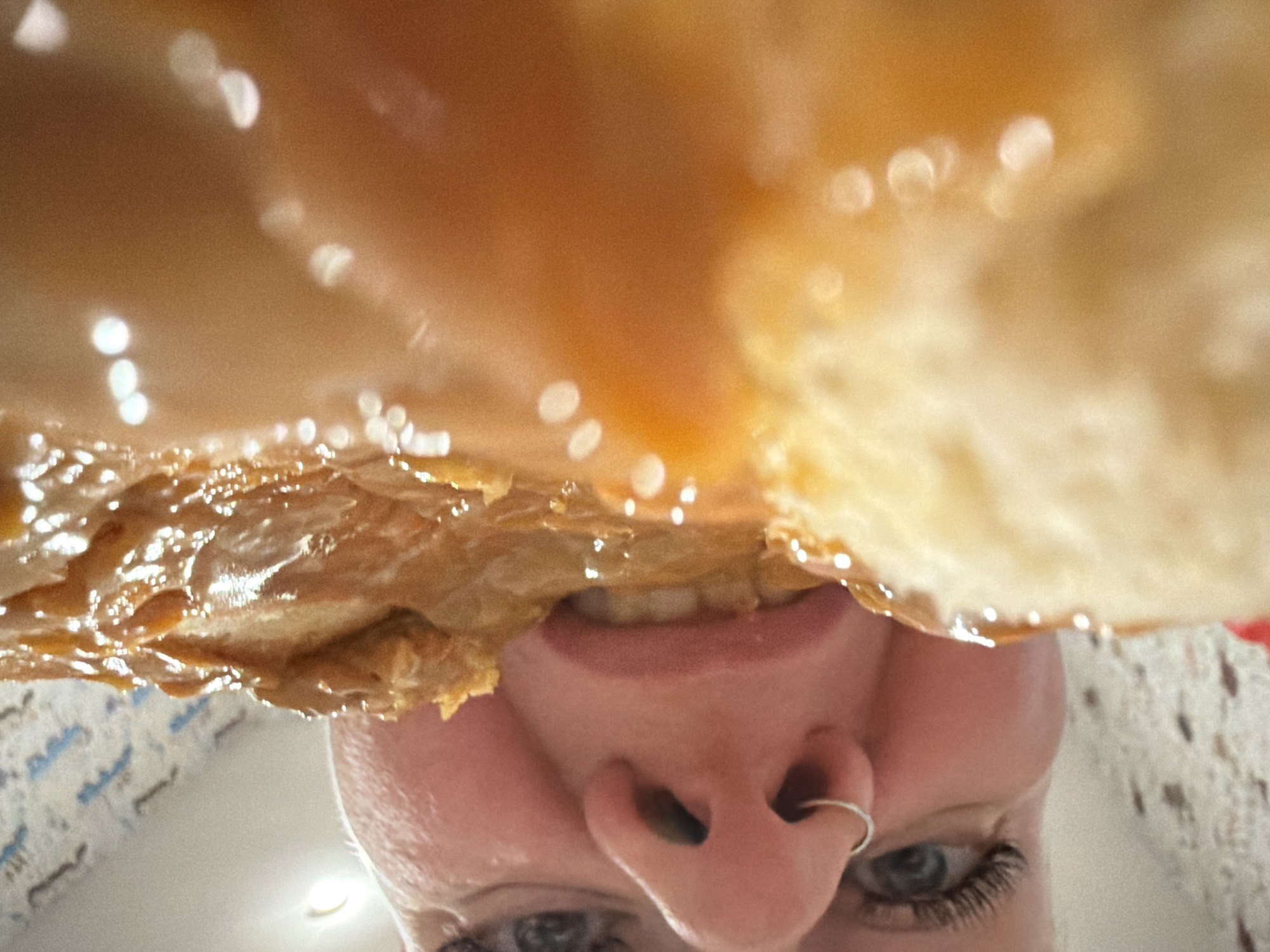 A person's face is seen looking up at a piece of food, possibly pastry or bread, covered in a glossy, brown glaze. The person has fair skin, visible teeth, a nose ring, and long eyelashes. The background is blurry and indistinct, with white and blue patterns suggesting a wall or other surface. The location is not determinable from this image.