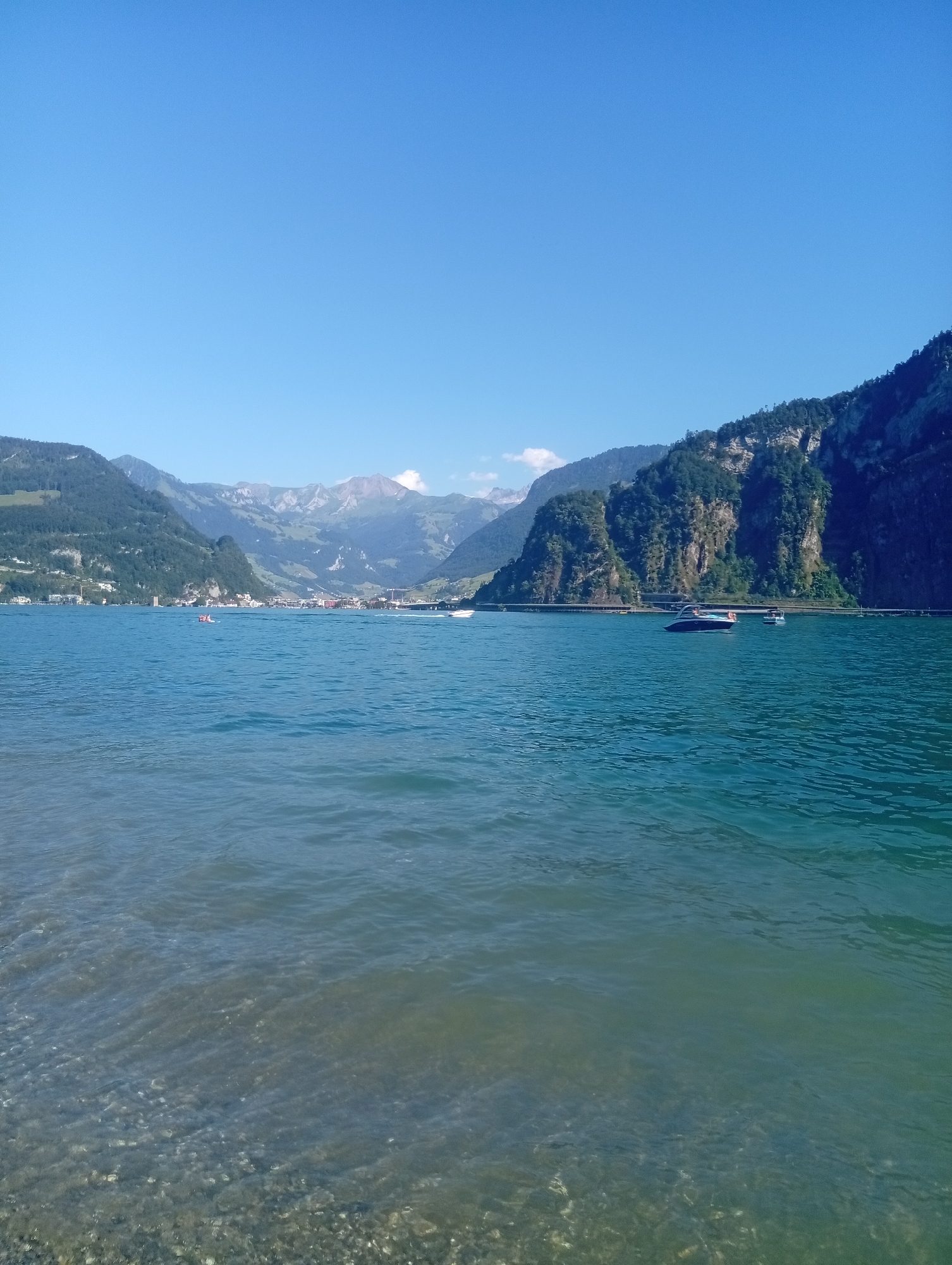 The image captures a tranquil scene on Lake Lucerne, looking towards the town of Hergiswil in the canton of Nidwalden, Switzerland. The foreground is dominated by the clear, shallow waters of the lake, revealing a pebble-strewn bottom. Gentle waves create ripples on the surface.
In the mid-ground, the expansive blue water extends towards the opposite shore. Several boats are visible: a small red inflatable craft with at least two people aboard is to the left of center, while a sleek motorboat with an orange life vest visible on its stern is positioned to the right of center. Another white boat is partially visible further to the right. A distinct, low bridge or causeway stretches across the water in the distance, connecting the shores.
The background is a dramatic panorama of mountains. The left side features a green, tree-covered slope leading up to rugged, layered mountains. In the center, a cluster of buildings representing the town of Hergiswil is nestled at the base of the mountains, with the higher peaks beyond showing patches of snow and rock. The right side of the background is dominated by a steep, dark, and craggy mountain face densely covered in trees. The sky above is a vast, clear, and brilliant blue. The overall impression is one of a serene summer day in a picturesque alpine setting.