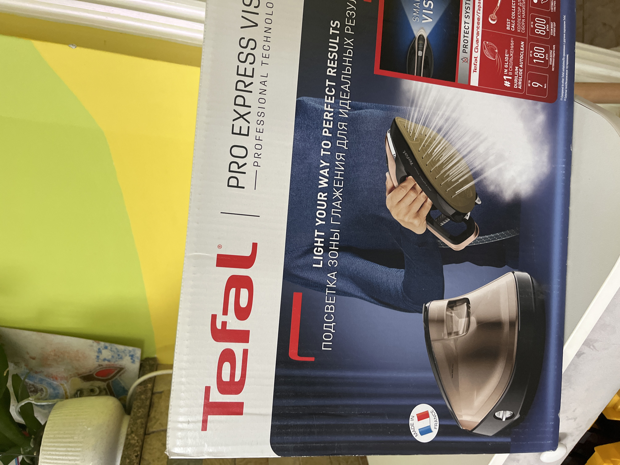 The image shows the front of a Tefal Pro Express Vision steam iron box. The box is predominantly white with a large red Tefal logo. Part of the box features an image of a person holding the steam iron, with steam emanating from it, and a close-up of the iron's soleplate. There is also an image of the steam generator base. Text on the box includes "LIGHT YOUR WAY TO PERFECT RESULTS", "ПОДСВЕТКА ЗОНЫ ГЛАЖЕНИЯ ДЛЯ ИДЕАЛЬНЫХ РЕЗУЛ", and details about the iron's features like "DURILIUM", "AIRGLIDE AUTOCLEAN", and steam output specifications. A "MADE IN FRANCE" sticker is visible on the left side of the box. The background of the box image is a dark blue, suggesting the product in use. The box itself is placed on a surface, possibly a shelf, with the top portion of the box showing. To the left and top of the box, a potted plant with white flowers is visible, along with some decorative items. The overall environment appears to be indoors, with a bright, possibly natural light source illuminating the scene from the top and right. The location being Onokivtsi, Ukraine, is not directly ascertainable from the visual elements of the image itself, but it is provided as contextual information.