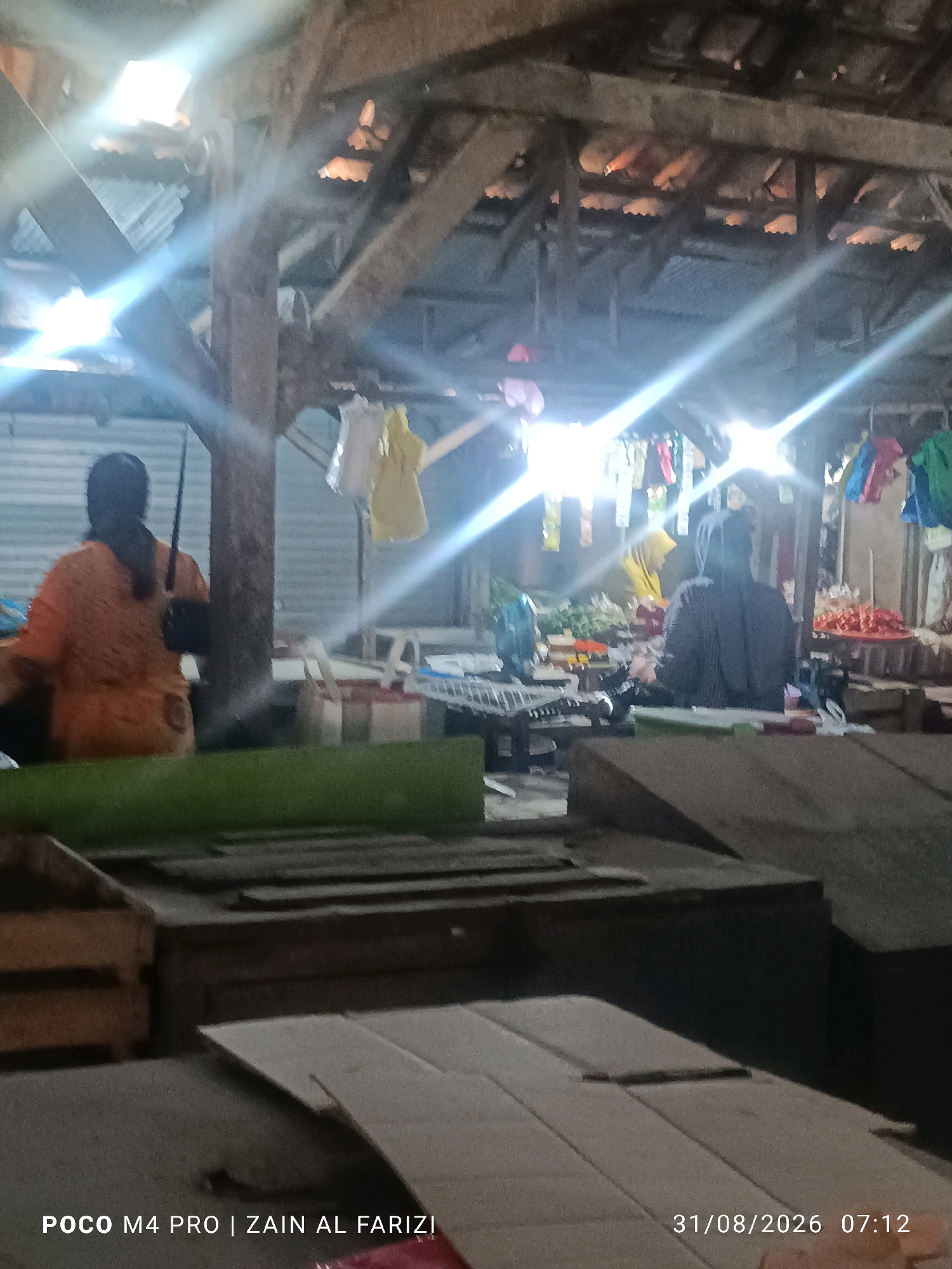 The image shows an indoor marketplace in Arosbaya, Indonesia, under a wooden beam roof structure with corrugated metal roofing visible in some areas. Bright lights are creating lens flares and illuminating the scene. In the foreground, there are wooden crates and what appears to be a tabletop covered with a green cloth. Further back, two individuals are visible. On the left, a person wearing an orange garment is seated, facing away from the camera, with a long dark object, possibly a stick or pole, held in their right hand. To the right of this person, there is another individual wearing a dark grey garment, also facing away from the camera. In the background, there are stalls displaying various items including what appear to be vegetables and possibly fruits, along with clothing hanging on display racks. Two light-colored bags are visible on a surface between the two seated individuals. The overall atmosphere suggests an active marketplace environment at a low-light time, possibly early morning or evening, given the artificial lighting and the timestamp indicating 07:12 on 31/08/2026.