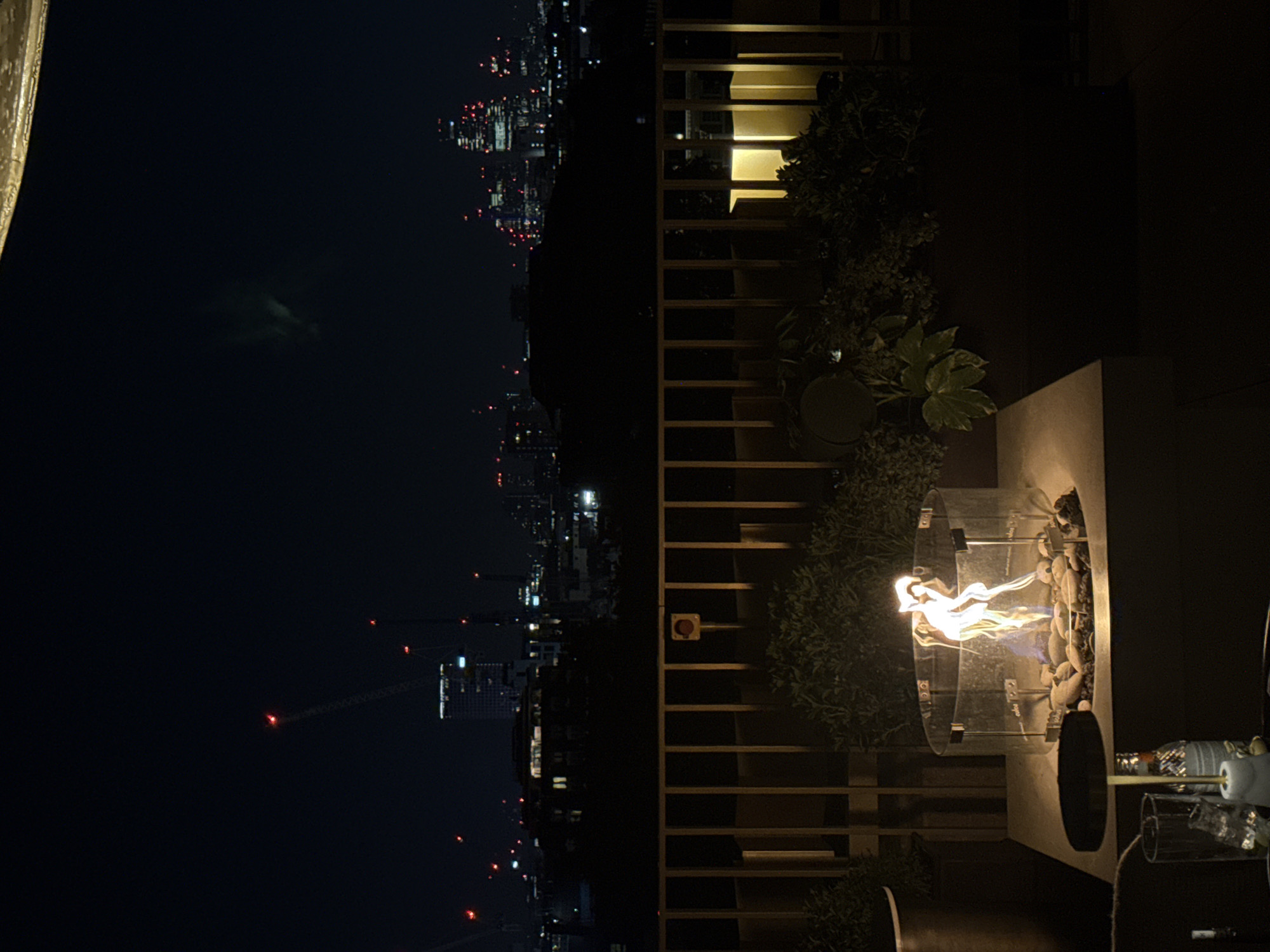 The image depicts a nighttime scene in London, United Kingdom, with a partial view of the city skyline visible in the background. A fire pit, containing flames and decorative rocks, is positioned in the foreground, along with glass and bottles to the side. Adjacent to the fire pit is a wooden structure resembling a balcony or partition, lined with plants. The environment is dark, with the primary light sources being the fire and the city lights in the distance.