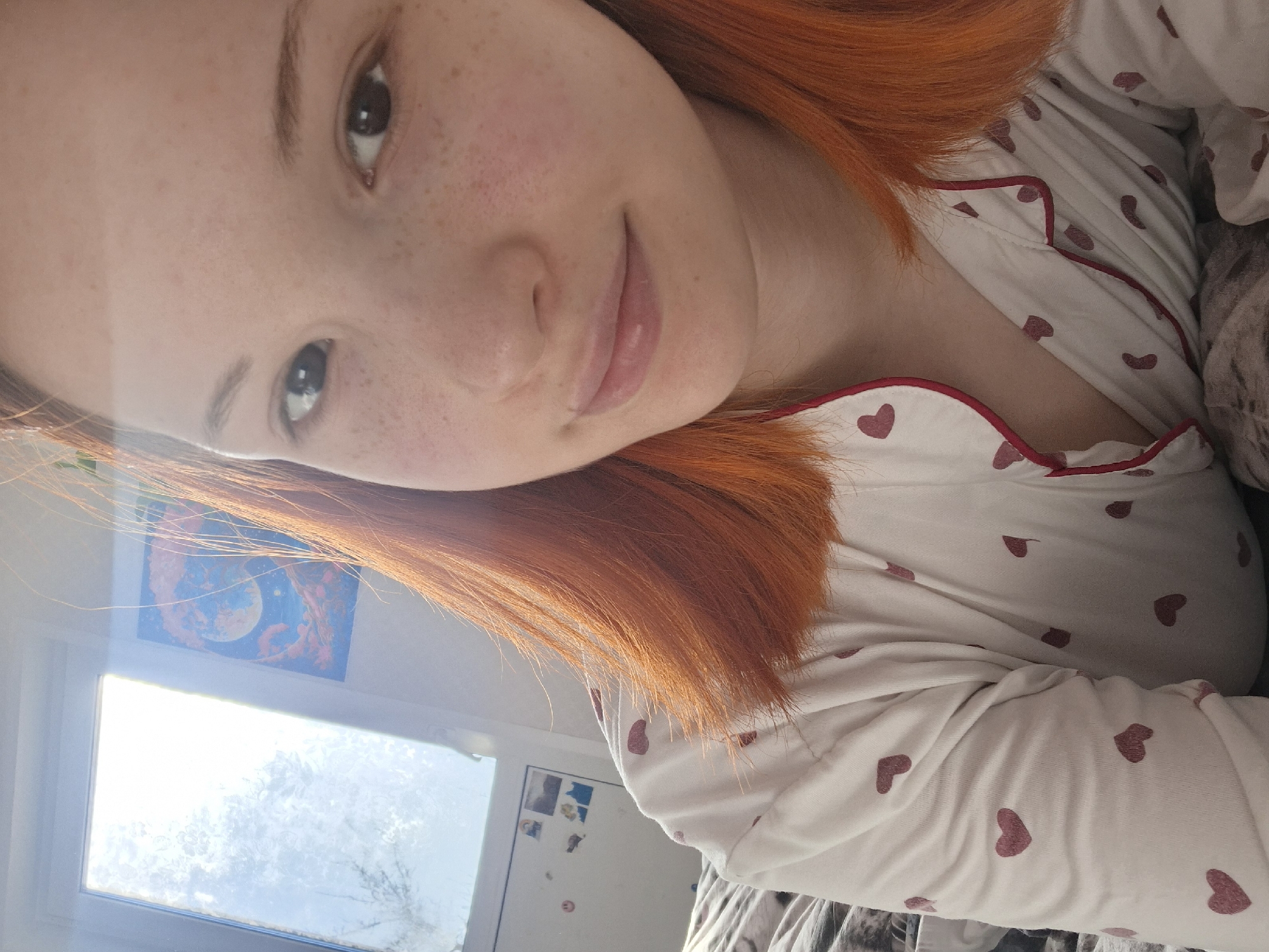 A young woman with red hair and freckles is lying down indoors. She is wearing white pajamas patterned with small red hearts and trimmed with red piping. Light streams in from a window in the lower left corner of the image. A blue and orange image of a planet is affixed to the wall above the window. A white refrigerator is also partially visible in the background. The location is in the city of Chaikovsky, Russia.