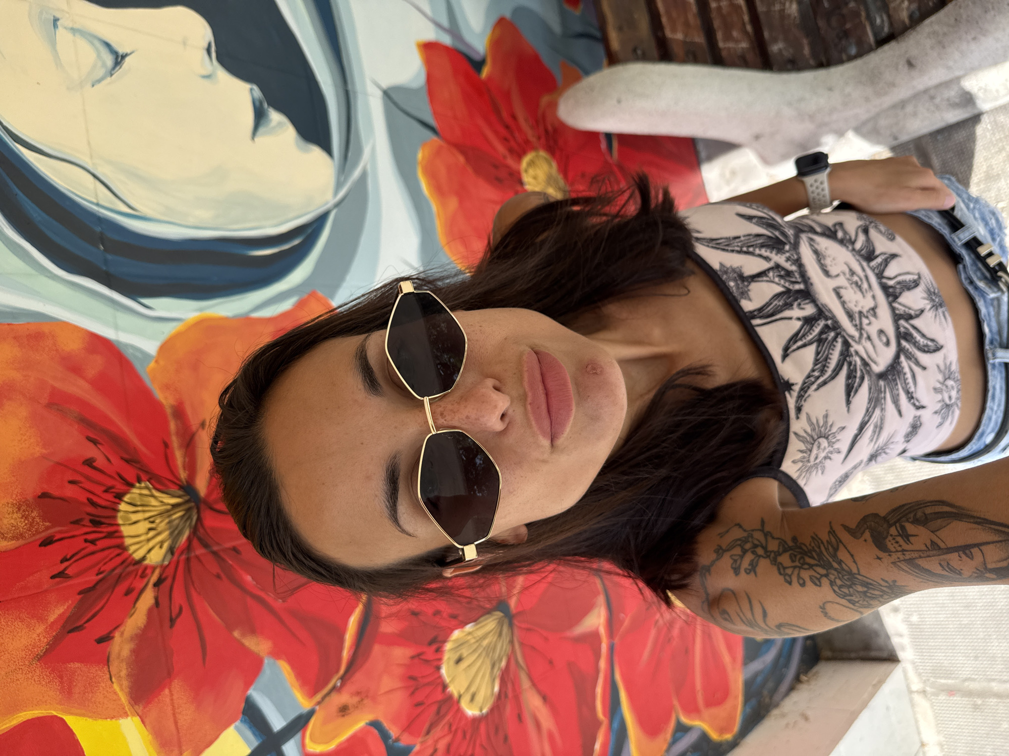This is a high-angle, medium shot of a woman with dark hair, wearing sunglasses and a patterned tank top, standing in front of a vibrant mural. The mural features large red poppies and a blue and white depiction of a woman's face. The woman is looking directly at the camera. Her left arm, visible in the frame, is covered in tattoos. She is also wearing denim shorts and a watch on her left wrist. A portion of a white, abstract sculpture or bench is visible to the right of the frame. The environment appears to be outdoors, with bright lighting suggesting daylight. The woman's posture and the framing of the shot suggest this is a selfie. The location is identified as Belene, Bulgaria.