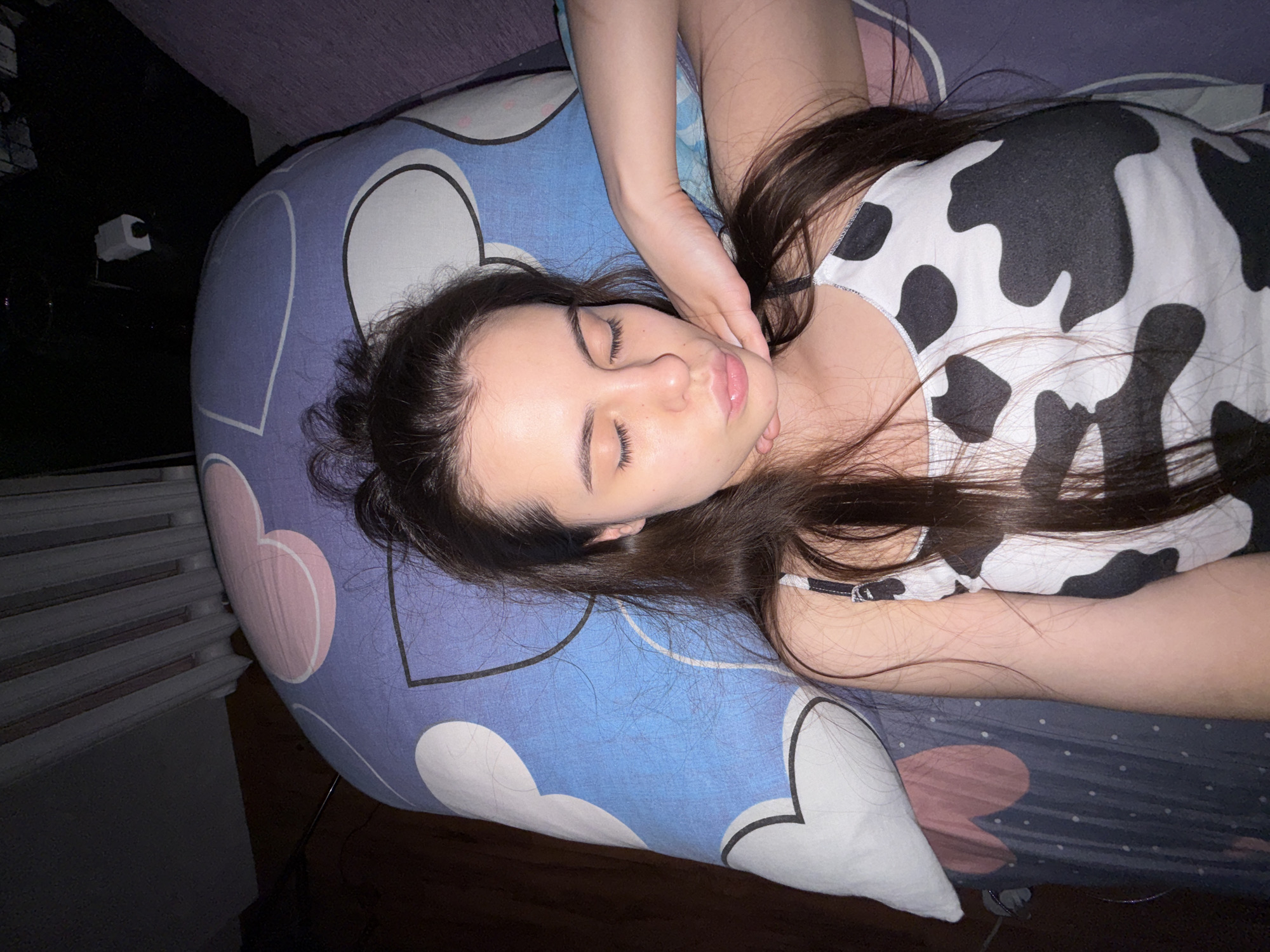 A young woman with long, dark hair is lying on a bed with a patterned blue and white pillowcase. She is wearing a white top with black cow spots. Her eyes are closed, and one of her hands is gently touching her neck. The setting appears to be a dimly lit bedroom, possibly in a private residence. It is impossible to verify if this image was taken in the city of Chaikovsky, Russia.