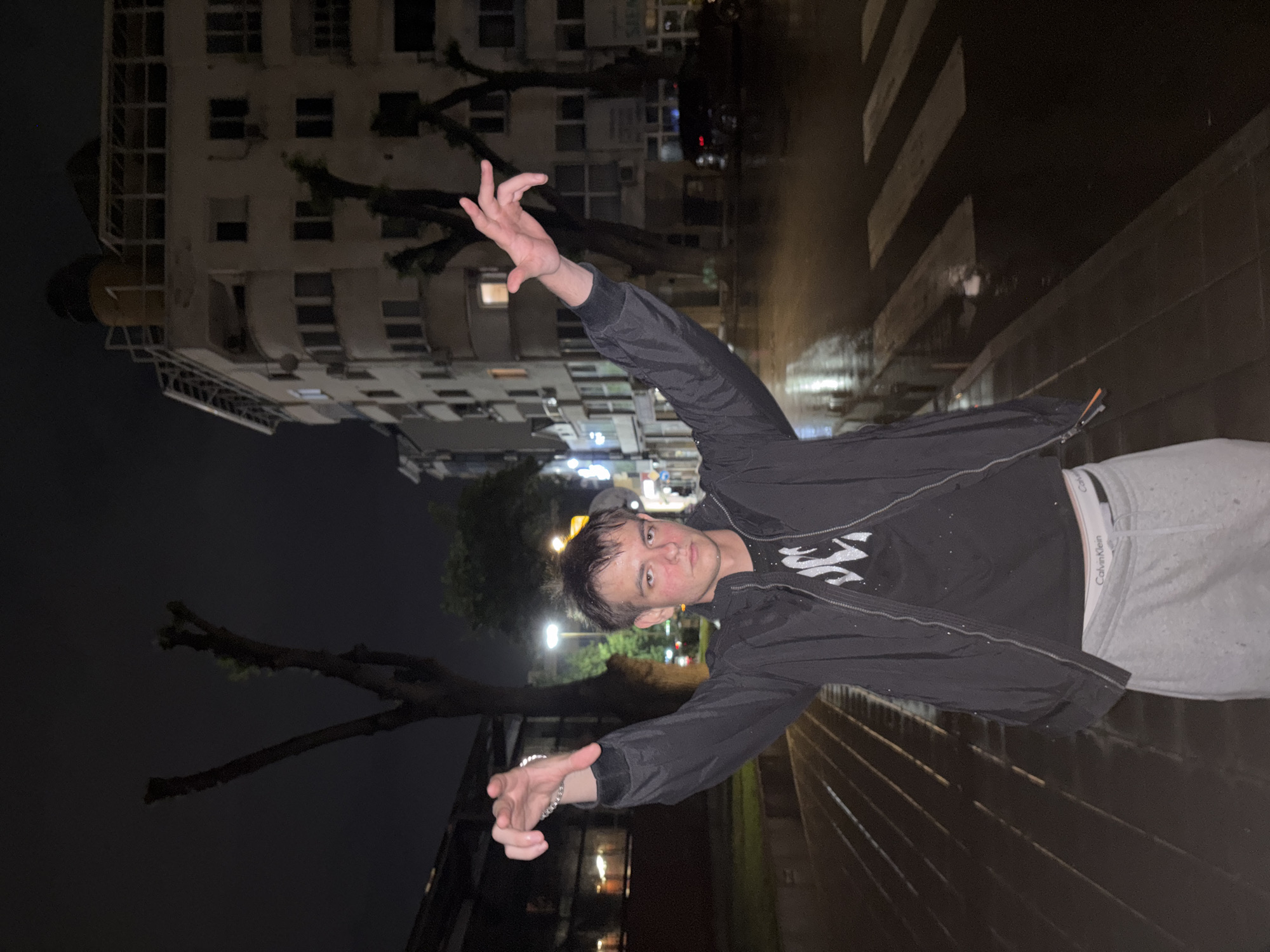 A young adult is standing on a street in Plovdiv, Bulgaria at night. The subject is wearing a dark jacket, a black T-shirt with a white design, and light-colored sweatpants with a visible Calvin Klein waistband. The subject has wet hair and is posed with arms outstretched, appearing to interact with the environment. The background shows a multi-story building, a street with visible crosswalk markings, and bare tree branches. The scene appears to be dimly lit, potentially after rain, as the ground reflects light.
