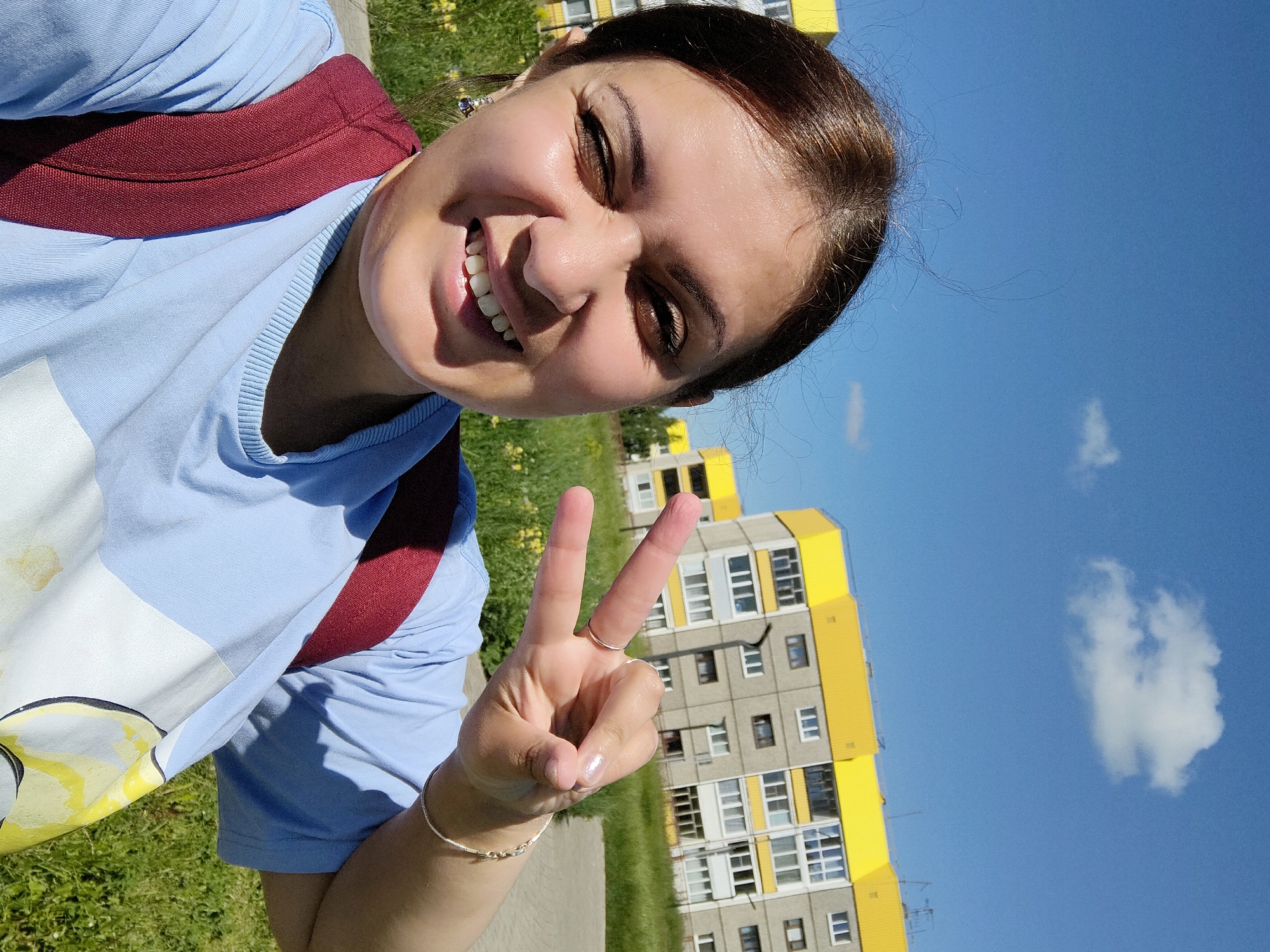 In an outdoor selfie, a young woman with brown hair and brown eyes is pictured smiling and winking while holding up two fingers in a peace sign. She is wearing a light blue t-shirt, a dark red backpack, and jewelry, including earrings and rings. Behind her, a modern apartment building with yellow accents and a clear blue sky with a few clouds are visible. The surrounding area appears to be a residential neighborhood with green grass and some trees. The image was taken in Markovsky, Russia.