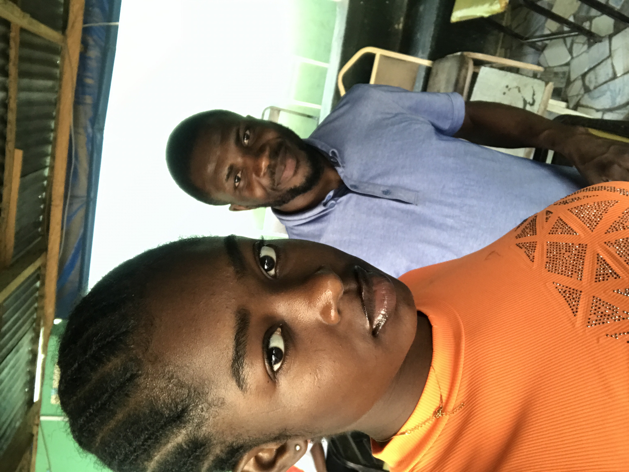 The image shows two individuals in an indoor setting, likely in the vicinity of New Stadium, Nigeria. The foreground features a young, dark-skinned woman with braided hair, wearing an orange top with small, diamond-shaped embellishments. She is looking directly at the camera. Behind her, there is a dark-skinned man with a closely-shaven beard, wearing a blue collared shirt and glancing at the camera with a slight smile. A metal roof structure and corrugated metal are visible in the background. Chairs are present behind the man, along with wall with stone-like texture. The lighting is bright, potentially from an overhead source.