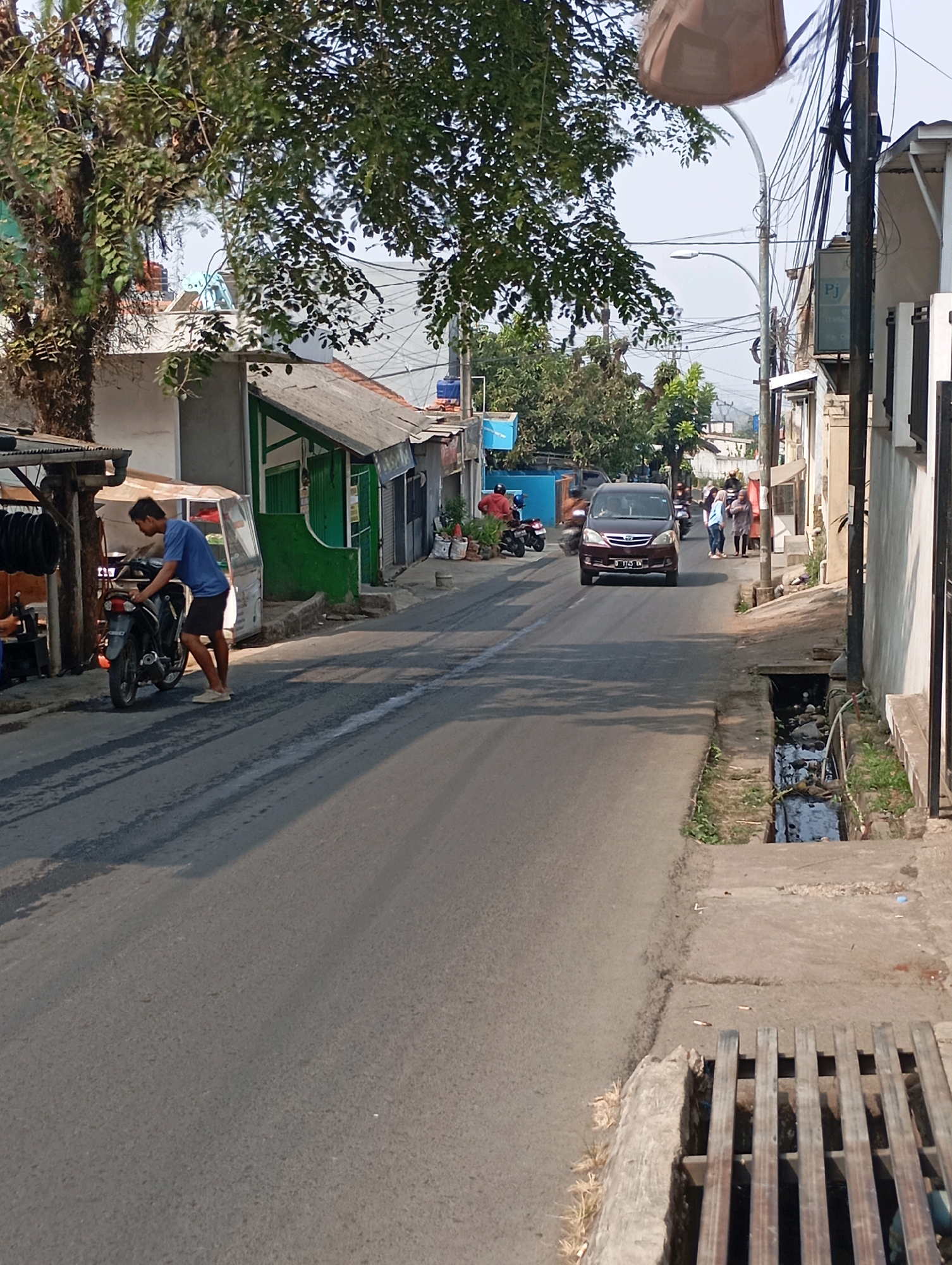 The image depicts a street scene in Cimahi, Indonesia, under a clear sky. In the foreground, a young man wearing a blue t-shirt and dark shorts is standing next to a black motorcycle, appearing to interact with it. The motorcycle is parked on the side of an asphalt road. Adjacent to the road is a concrete sidewalk and a drainage ditch filled with water and debris. Buildings line the street, some appearing to be shops or small residences with green and grey facades. Further down the road, a dark minivan is driving away from the camera, and several pedestrians are visible on the sidewalk. Overhead, numerous electrical wires are strung between utility poles, a common sight in urban Indonesian settings. The environment suggests a typical residential and commercial street in a city.