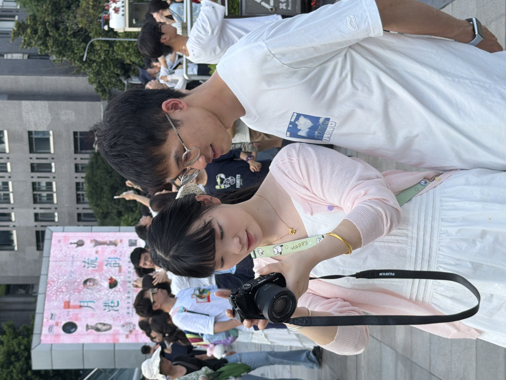 In the Yuzhong District of China, a young woman with dark hair styled in bangs, wearing a white dress and a light pink cardigan, is holding a black camera with a Fujifilm strap. She is looking down at the camera, seemingly focused on its display. Beside her, a young man with short dark hair and glasses, wearing a white t-shirt with a blue graphic and a white long-sleeved shirt underneath, is looking at her and smiling. They are standing in an outdoor public space, likely a street or plaza, with a large digital billboard displaying pink graphics and Chinese text in the background. Other people, some holding phones aloft, are visible in the crowded scene, suggesting an event or gathering. The environment appears to be urban, with a large building and trees in the background. The lighting suggests it is daytime.