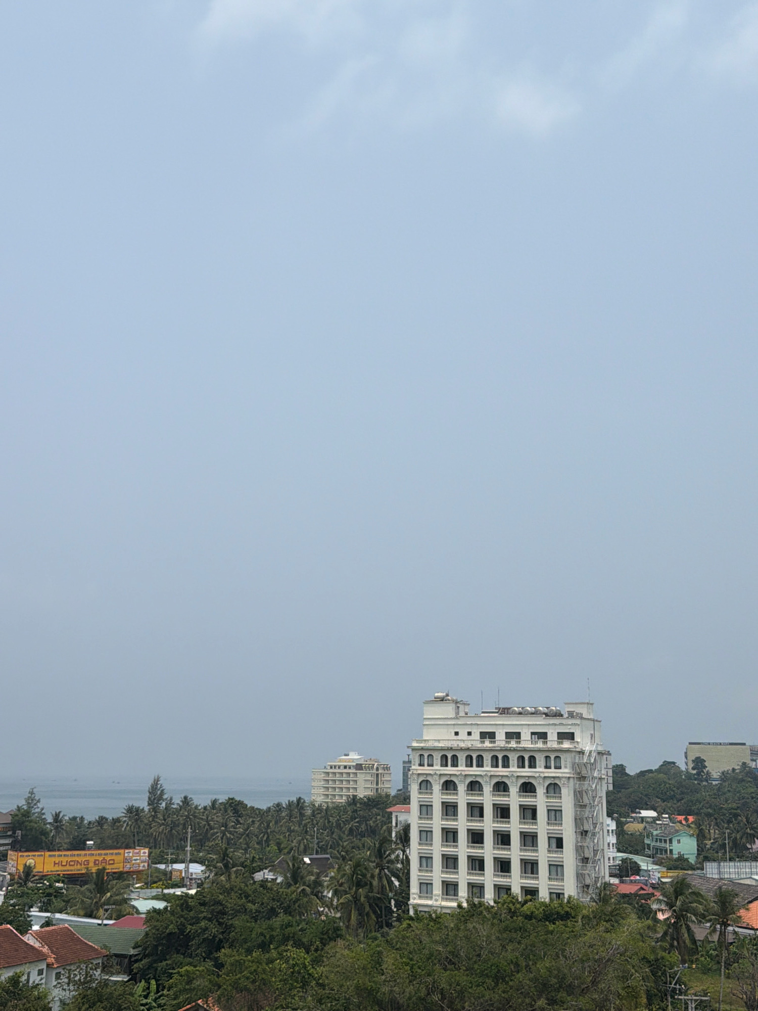 The image shows a cityscape in Phú Quốc, Vietnam, with a focus on a tall, white, multi-story building with arched windows and a fire escape. The building is surrounded by lush green trees and other smaller buildings. In the background, the ocean is visible, with a hazy sky above. A large billboard with Vietnamese text is also present in the foreground, advertising "HUONG DAO". The scene appears to be taken from a high vantage point, offering a panoramic view of the area.