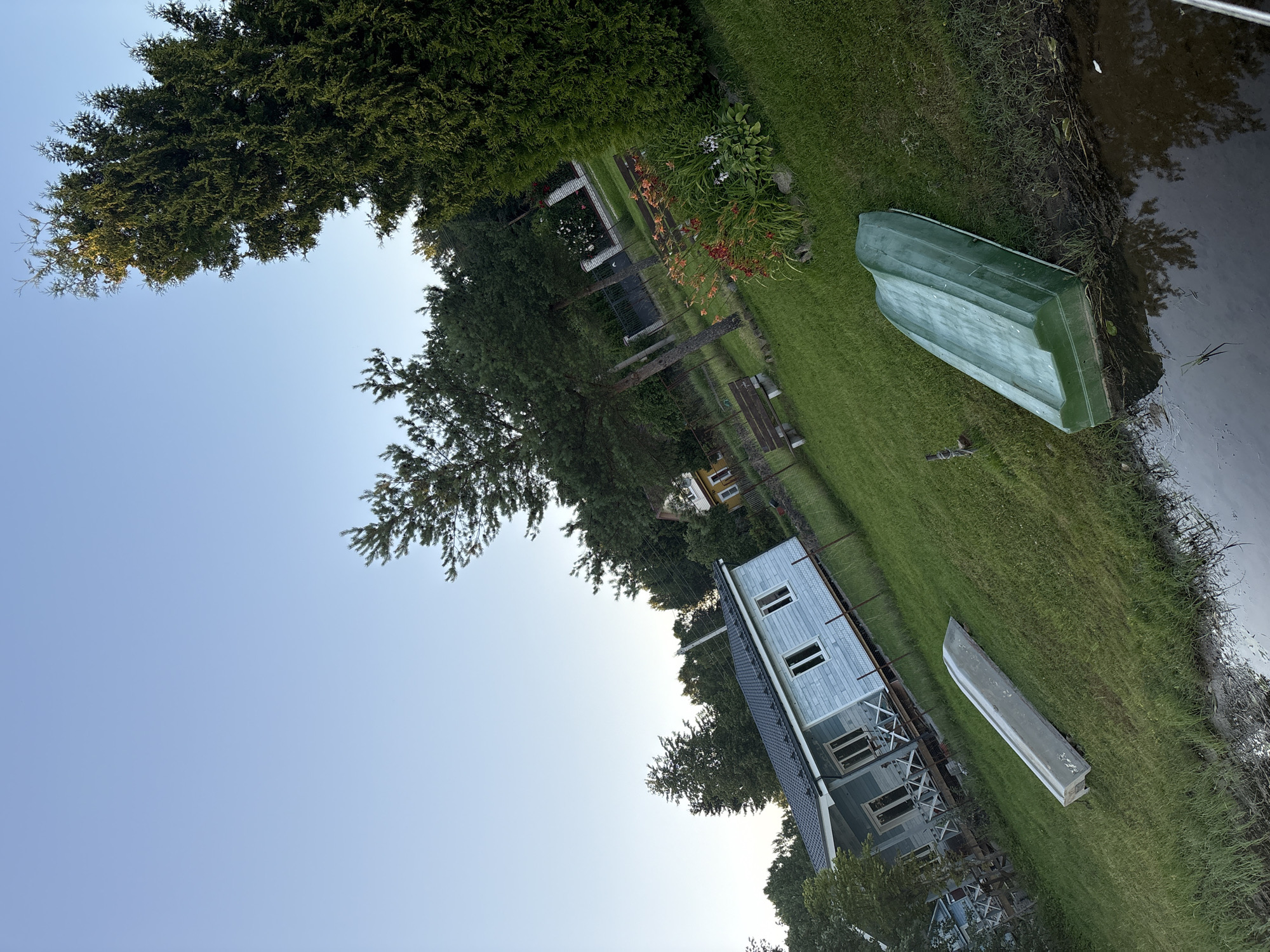 In Chikino, Russia, an outdoor scene unfolds under a clear sky. Two boats are present on a grassy slope leading down to a body of water. One is a silver boat, resting upside down, and the other is a green boat, also upside down, situated closer to the water's edge. A light blue house with white trim and a dark roof stands on the left side of the frame. Beyond the house, trees and foliage are visible, with a hint of a yellow house further in the background. The water in the foreground reflects the surrounding trees and sky. The overall impression is of a tranquil, rural setting.