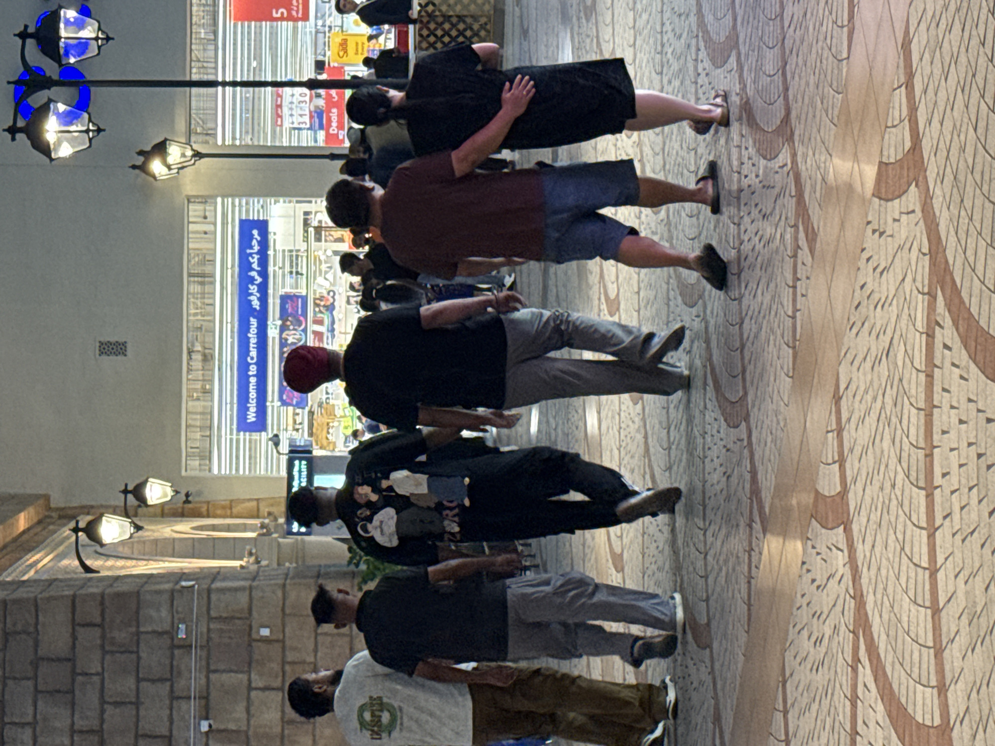 The image shows a group of people walking in an outdoor, pedestrianized area, likely a shopping district or promenade in Dubai, United Arab Emirates. The architecture suggests a modern commercial setting with a large brick building on the left and the facade of a Carrefour supermarket with illuminated signage in the background. Streetlights with a decorative blue light are visible, along with lampposts that cast a warm glow. The ground is paved with patterned tiles.
The foreground focuses on the backs of six individuals walking away from the camera. From left to right:
1. A person wearing a light-colored t-shirt with text and dark brown trousers.
2. A person wearing a black short-sleeved shirt and gray cargo pants.
3. A person wearing a black t-shirt with a graphic print on the back, black trousers, and dark shoes.
4. A person wearing a maroon t-shirt and blue shorts.
5. A person wearing a black dress or long top, with their arm around the person in front of them.
6. The back of a person's head, wearing a red turban, is visible, positioned further back in the group.
Other people are visible in the background, suggesting a public space with moderate foot traffic. The overall atmosphere appears casual and urban, illuminated by artificial lights indicating it's likely evening or nighttime. The Carrefour signage "Welcome to Carrefour" is prominently displayed in both English and Arabic.