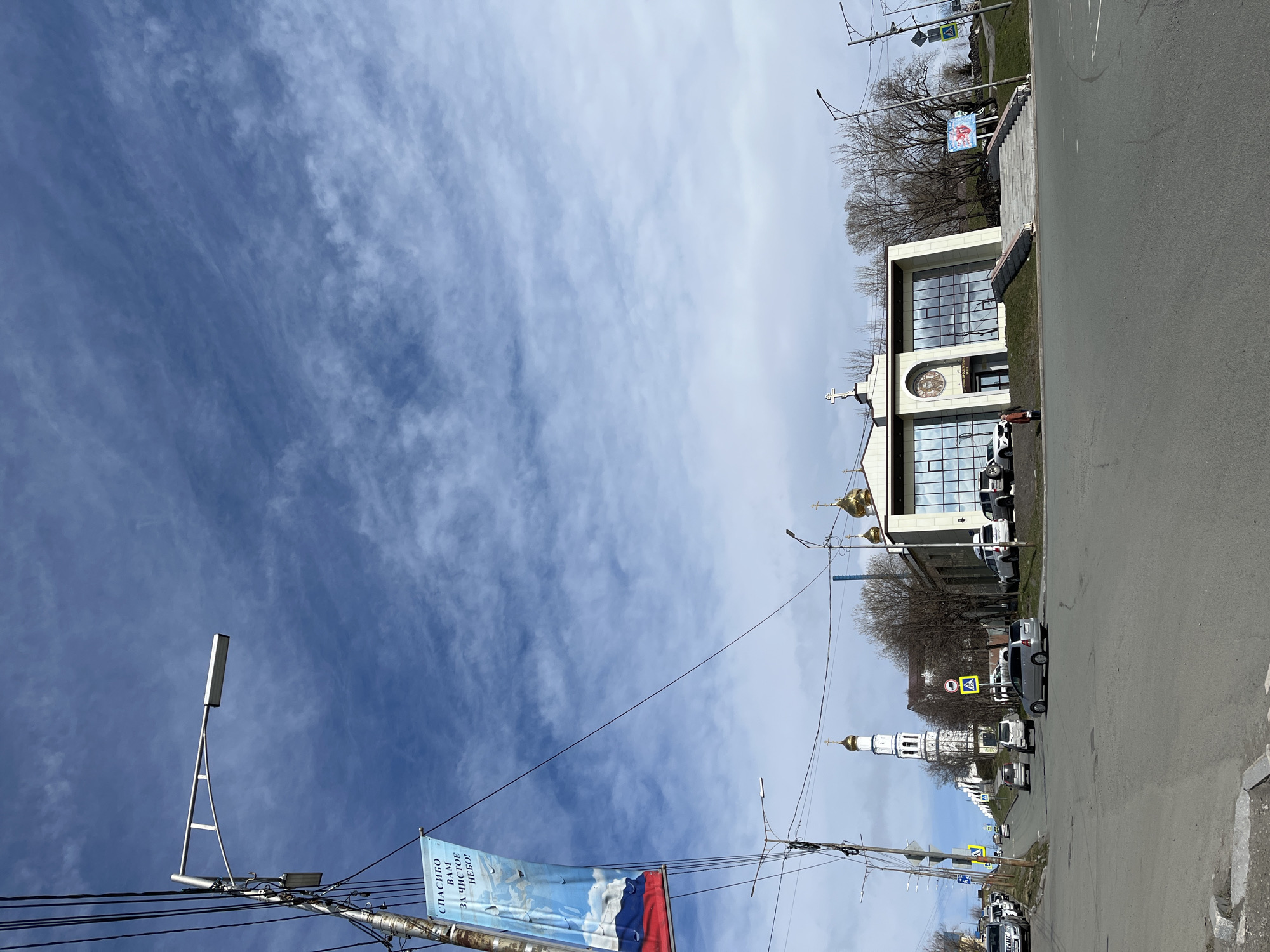 The image shows a street scene in Petropavlovsk-Kamchatsky, Russia. The sky is predominantly blue with wispy clouds. On the right side of the frame, a modern-style church with a large clock and a cross on top is visible. It has two floors and large windows. In front of the church, there are several parked cars. Further down the street, a traditional white church with a golden dome is visible. Streetlights and power lines run along the left side of the road. A banner with the Russian flag and text "Спасибо Вам за чистое небо!" (Thank you for the clear sky!) is attached to a utility pole. The road is asphalt and appears to be in moderate condition. The surrounding area has some trees and greenery, though the trees are mostly bare. Several road signs are posted along the street.