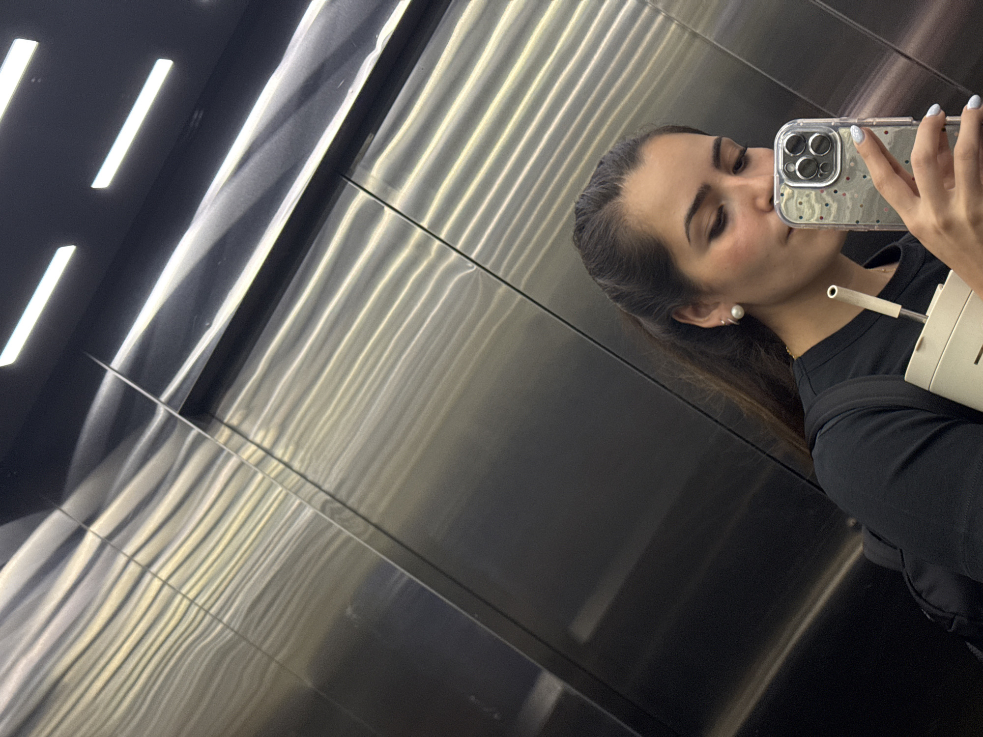 A young woman takes a selfie in an elevator with brushed metal walls. The elevator has horizontal fluorescent lighting. She holds a phone with a clear, polka-dotted case up to her face, angled so her face is visible. She is wearing a black top, a small pearl earring, and appears to have a beverage container and straw in her other hand. The location in San Bernardino Tlaxcalancingo, Mexico is not identifiable from the visual information.