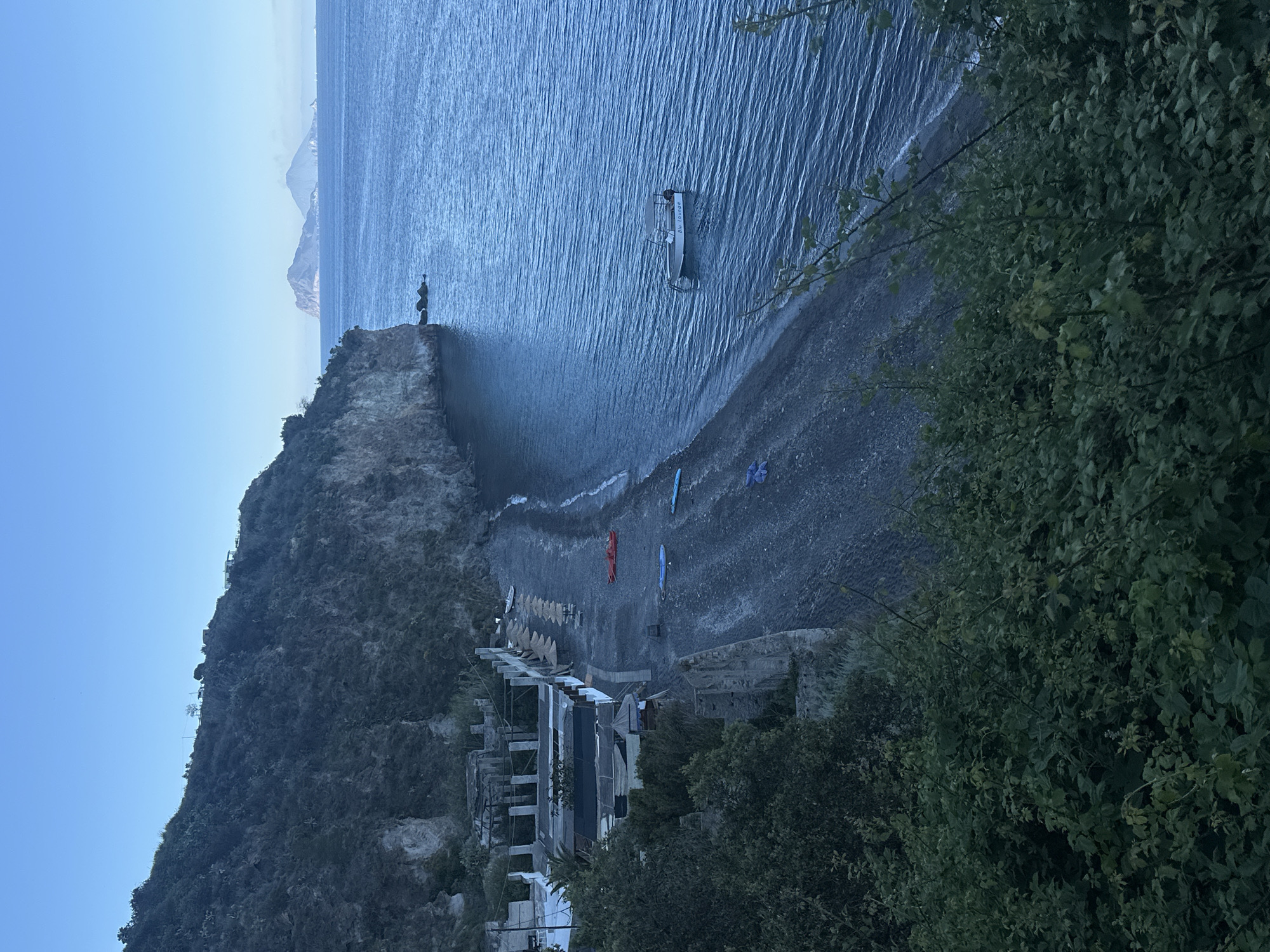 This image shows a view of Spiaggia Praia di Vinci on the island of Lipari, Italy. The photograph is taken from an elevated position, likely overlooking the beach from the hillside.
The scene includes:
* **Landscape:** A dark beach with a rocky cliff face extending into the sea. In the distance, there are hints of mountainous land.
* **Sea:** A calm, blue body of water. A small boat or platform is visible, anchored or floating just off the coast.
* **Beach:** The beach has a dark, pebble or rock composition. Various objects are scattered along the beach.
* **Structures:** Some buildings or shelters are partially visible along the beach. The architecture appears rudimentary.
* **Vegetation:** Dense greenery, likely trees and shrubs, frames parts of the scene, suggesting the vantage point is from within or near a wooded area.
* **Objects:** At least 3-4 paddle boards are visible on the beach.
The overall impression is of a relatively secluded, undeveloped beach location. The lighting suggests either dawn or dusk. There are no obvious signs of human activity in the immediate view.