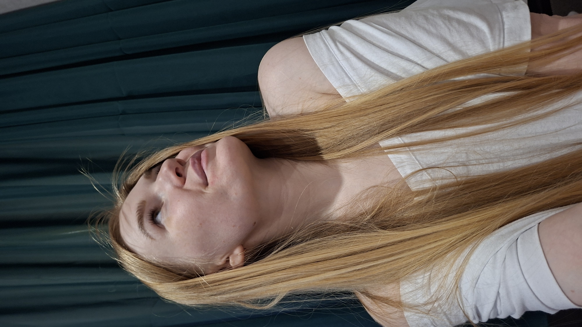 A young woman with fair skin and long, straight, blonde hair is lying on a dark teal surface with horizontal lines. She is wearing a white t-shirt. The location is not identifiable as Chaikovsky, Russia from the given image.