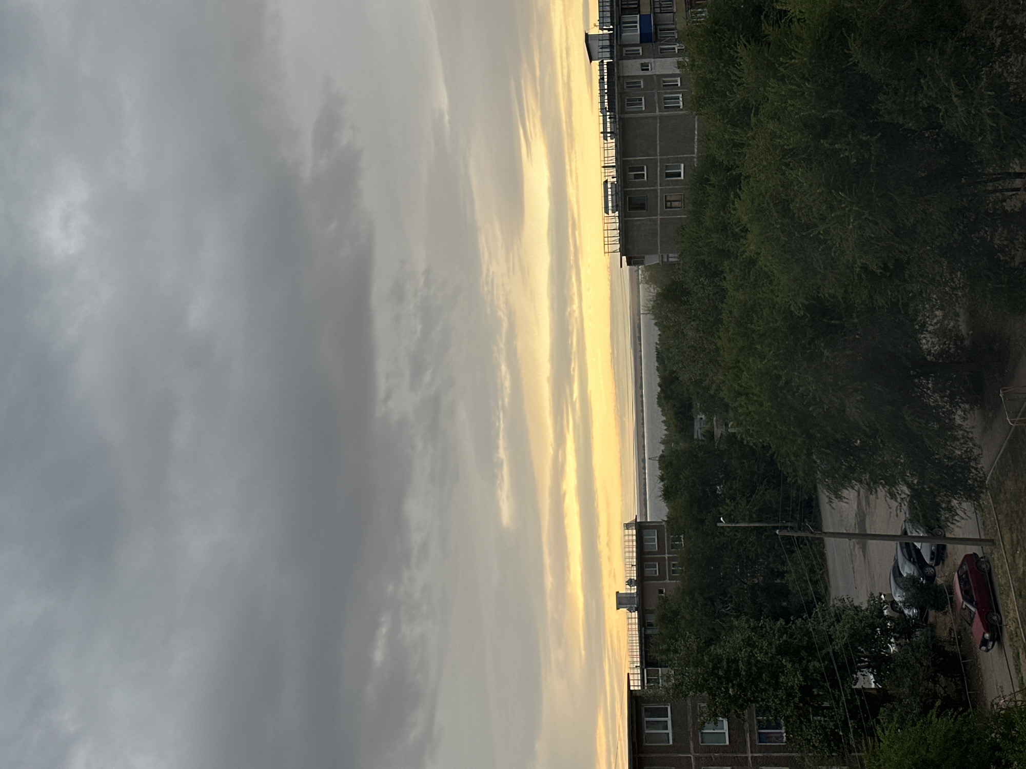 A wide shot captures a cloudy sky with bands of yellow light near the horizon, suggesting either sunrise or sunset. Below the sky, a body of water is visible, with a distant shoreline. In the foreground, the tops of apartment buildings and lush green trees are prominent. Cars are parked on a street in front of the buildings, and a single red car is parked closer to the viewer. The scene appears to be a residential area in the city of Energetik, Russia.