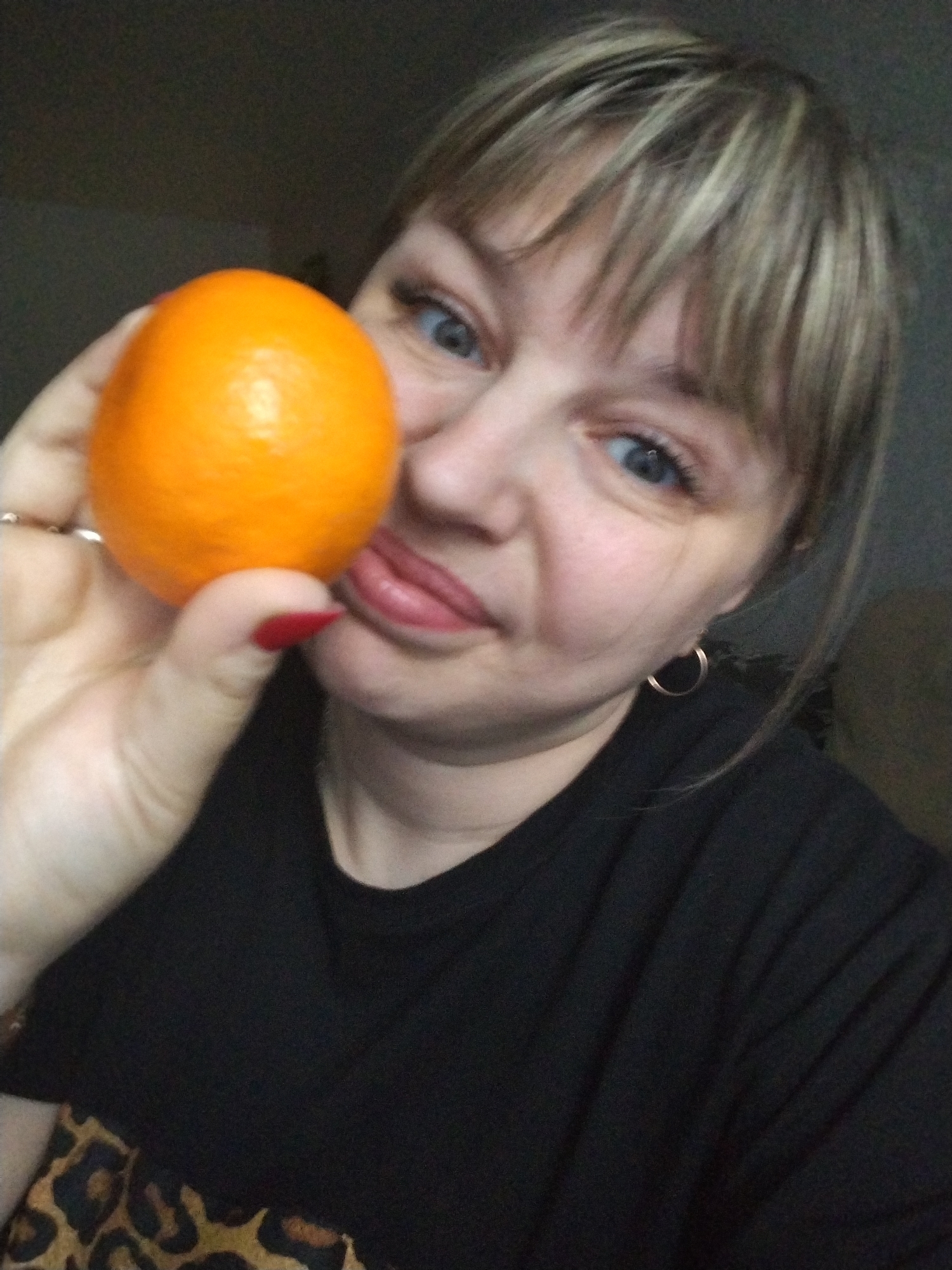 A woman holds an orange up to her face in a selfie-style photograph. The woman has fair skin, blue eyes, and blonde hair with bangs. She wears a black shirt and gold hoop earrings. Her fingernails are painted red. The orange is bright and smooth, held in her left hand. Part of a leopard print garment is visible at the bottom left of the image. The photograph appears to have been taken indoors with soft lighting. There is no clear indication of Yaroslavl, Russia.