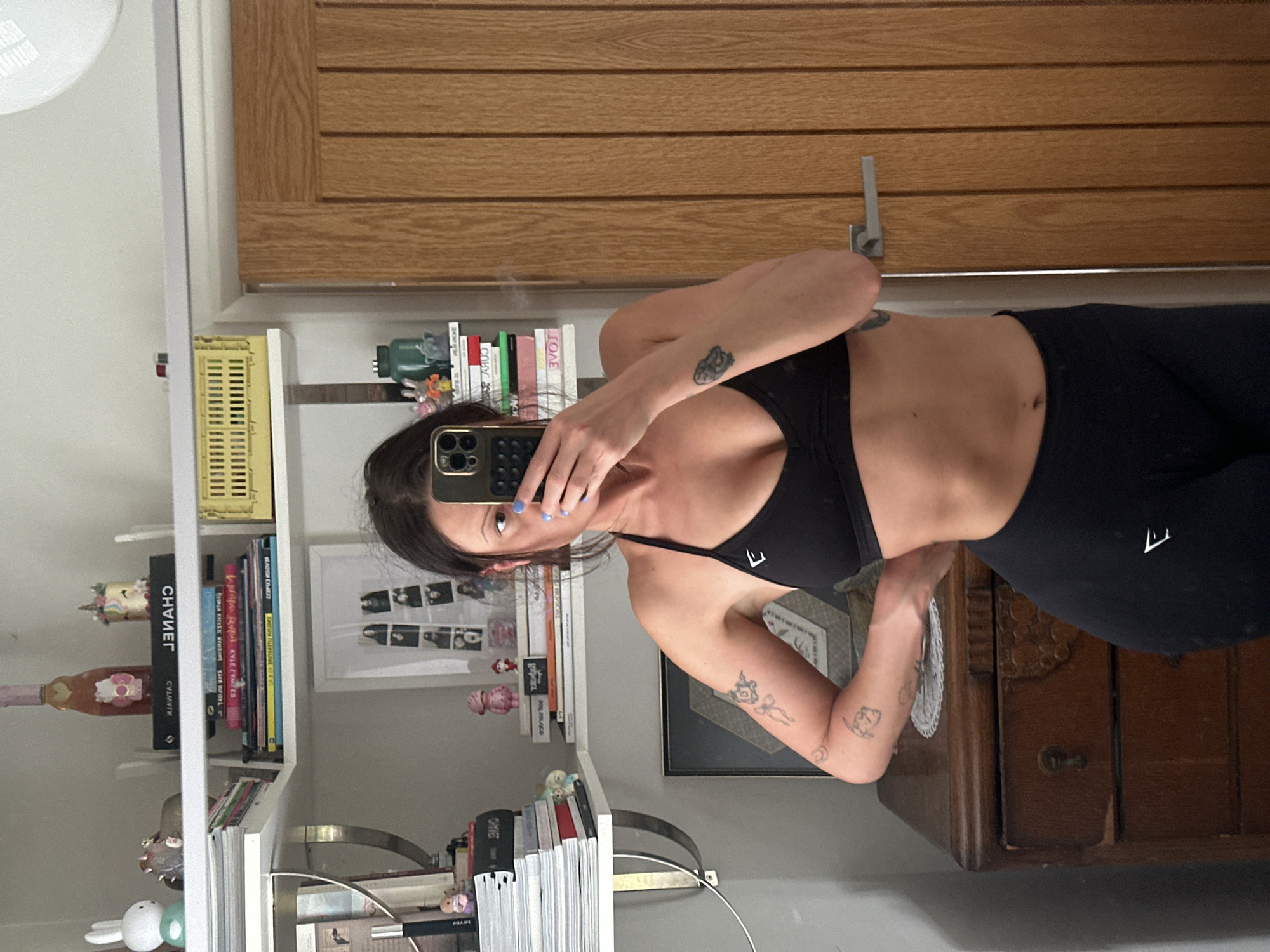 The image shows a woman taking a selfie in a mirror. She is wearing a black sports bra and black leggings. The background includes a bookshelf with books and various decorative items, a wooden door, and a wooden dresser. There is no evidence to place this image in Tunbridge Wells, United Kingdom.