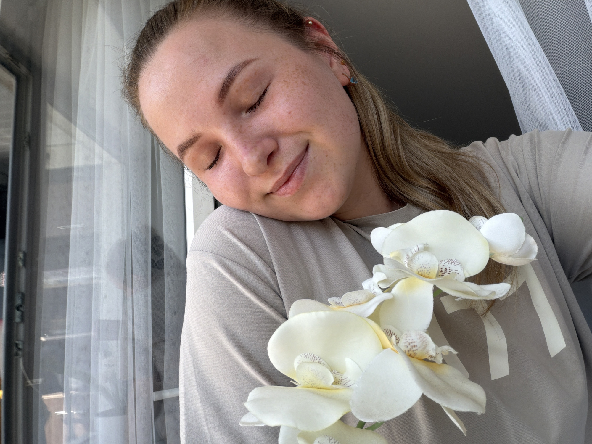 A young woman with blonde hair tied back and freckles on her face is holding a white orchid to her cheek. She is wearing a light brown t-shirt and has her eyes closed, with a slight smile on her face. The background consists of sheer white curtains and a dark wall, suggesting an indoor setting near a window. The image is taken from a high angle, looking down at the woman. The location is not specified in the image or its context.