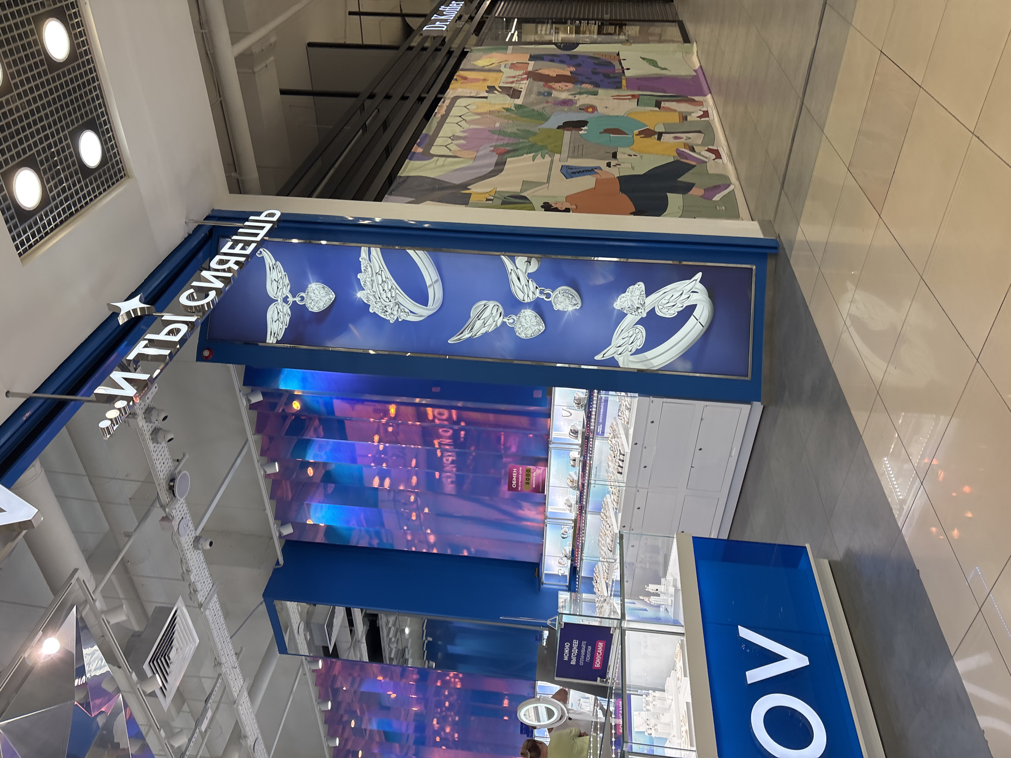 The image shows the interior of a jewelry store with a prominent blue and white color scheme. A large banner with a white inscription, which reads "...И ТЫ СИЯЕШЬ" (And you shine), hangs on a blue pillar. Below the inscription, there are images of rings and heart-shaped pendants with wings. In the foreground, a blue display case with the white letters "OV" is visible, possibly part of the store's branding. To the right, a store with a colorful mural depicting people shopping is adjacent. The store is located in Naberezhnye Chelny, Russia.