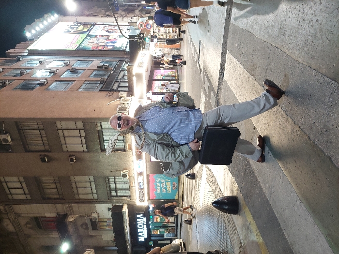 A man stands on a crosswalk holding a black briefcase. He's wearing light khaki pants, a light blue shirt, a beige jacket, and sunglasses. A patterned scarf is around his neck. Buildings line the street with stores below and apartments above. Streetlights and store signs illuminate the scene. The photo is taken at night and is slightly tilted to the left. Other people and parked cars are visible in the background.