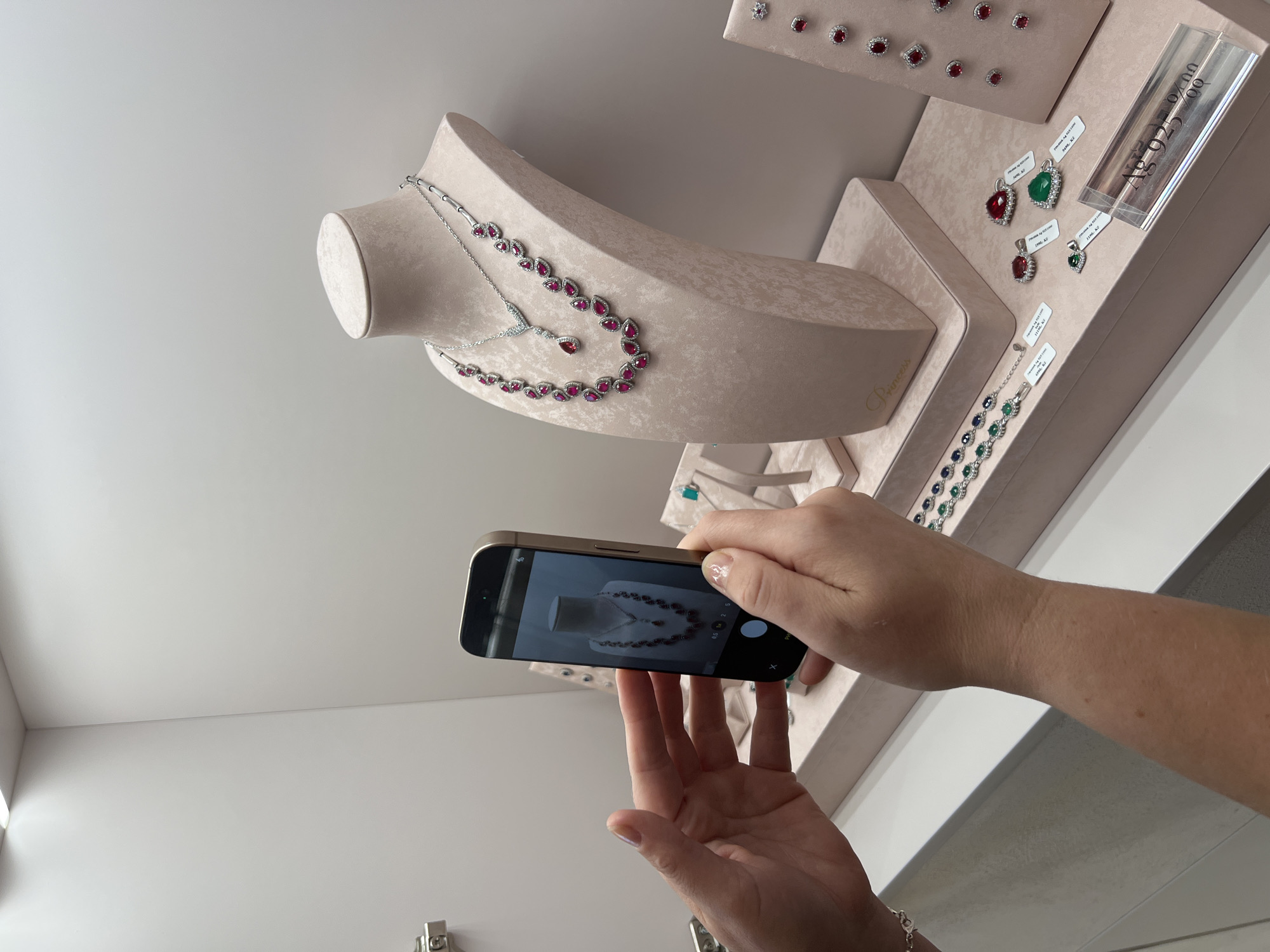 In an indoor retail setting, a person's hands hold a smartphone and are positioned to take a photograph of jewelry displayed on mannequins and stands. The background consists of white, minimalist walls and shelving. On a pink, velvet-covered mannequin bust, a silver necklace adorned with teardrop-shaped red gemstones is prominently displayed. To the right, a pink display stand showcases multiple pairs of earrings, each featuring red gemstones. In the foreground, a pink velvet tray holds loose pendants with red and green gemstones, as well as a bracelet with blue and green stones. A clear price tag with "Ag 925 925 00" is visible on the right. The overall environment appears to be a jewelry store or a display area within a department store.