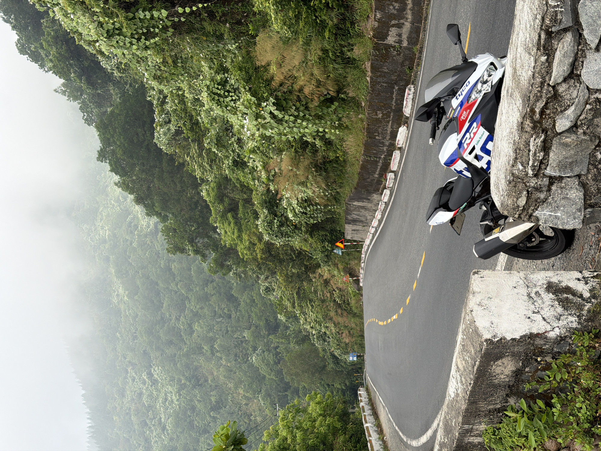 A motorcycle is parked on a winding road in Đà Nẵng, Vietnam. The motorcycle is mostly white with red and blue accents. The road is black asphalt with yellow dashed lines indicating a curve. The scene overlooks a lush, green mountainous area with some fog or mist in the distance. The road is bordered by concrete barriers and natural stone walls. The motorcycle is partially leaning against the stone wall.