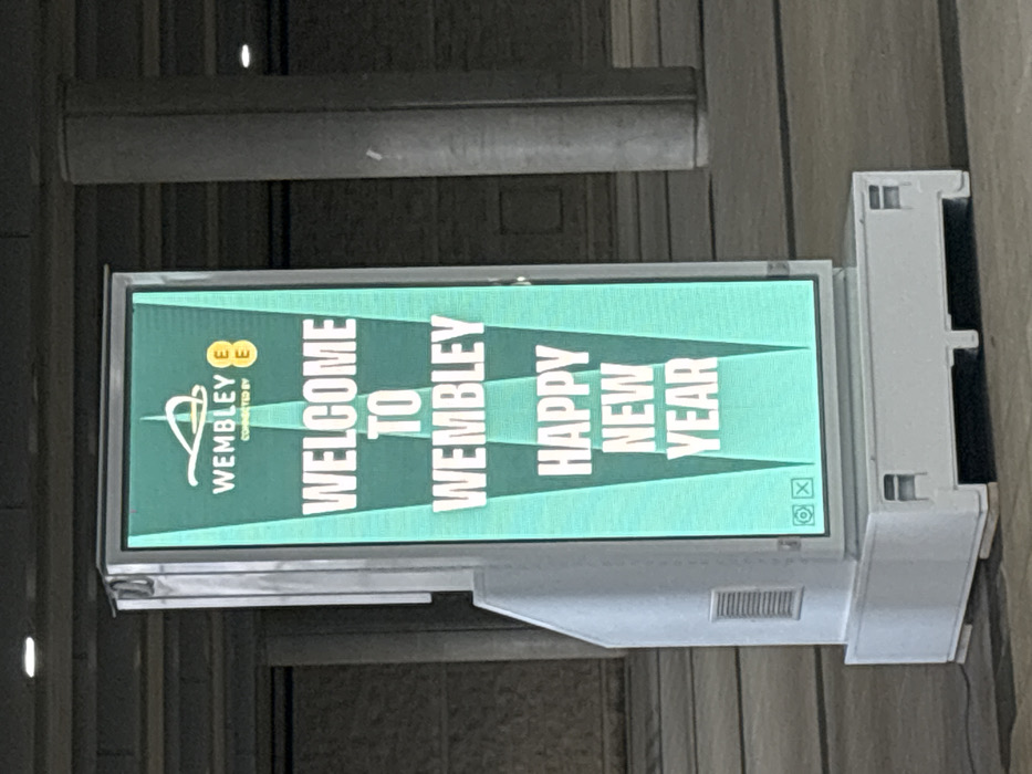 A brightly lit, vertically oriented digital display screen is the central subject, featuring a vibrant green background with prominent white text and graphics. The screen is mounted within a light gray frame, which appears to be part of a larger fixture, visible with a ventilation grille on its lower right side.
The display shows a welcoming message. In the upper left corner, the Wembley logo, an abstract 'W' design, is visible, accompanied by the words "CONNECTED BY EE" and the orange EE logo. The main message, stacked vertically and readable from top to bottom, states "WELCOME TO WEMBLEY HAPPY NEW YEAR." The text is large, bold, and white, set against the green background, with subtle lighter green triangular shapes extending from the text. In the bottom right corner of the screen, small icons, including a camera symbol and a square, are visible.
The screen is set against a dark, indistinct background, likely a wall or structural element, suggesting an indoor or covered public area. The lighting in the background is dim compared to the illuminated screen. The overall scene indicates a festive and welcoming atmosphere, consistent with a public venue in Wembley, United Kingdom, around the New Year period.