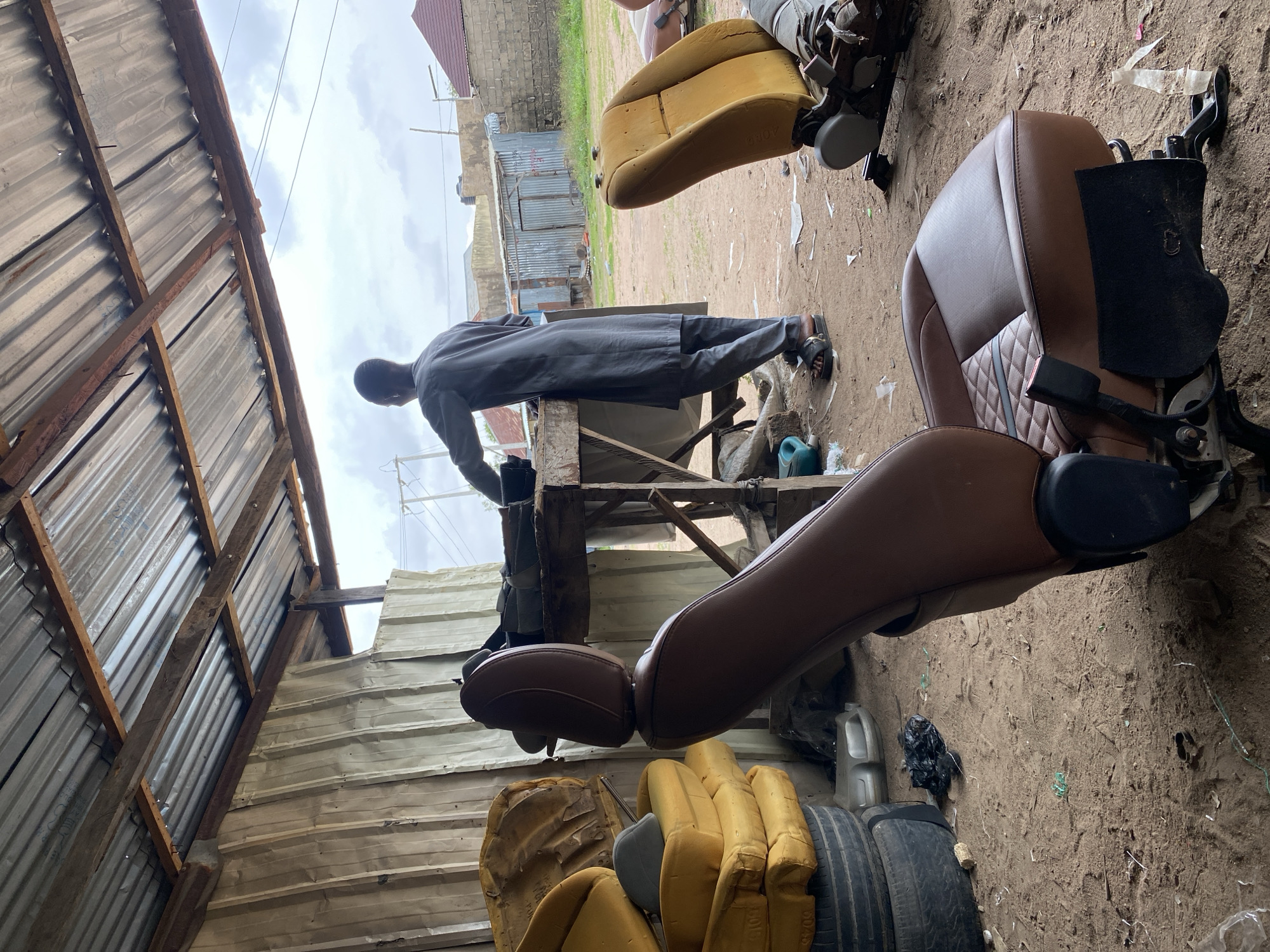 The image depicts an outdoor workshop in Jalingo, Nigeria, likely dedicated to automotive upholstery. A man, dressed in traditional grey attire, stands with his back to the viewer, engaged in work at a wooden table. Scattered around the workshop are various car seat components, including disassembled yellow and brown car seats, some appearing worn and others in better condition. A brown car seat is prominently featured in the foreground, showcasing quilted detailing on its backrest. The ground is covered in dirt and debris, with discarded materials such as tires, plastic bags, and scrap metal evident. The workshop is partially covered by a corrugated metal roof supported by wooden beams, suggesting a rudimentary construction. In the background, glimpses of residential buildings and the sky can be seen, indicating an urban or semi-urban setting within Jalingo.