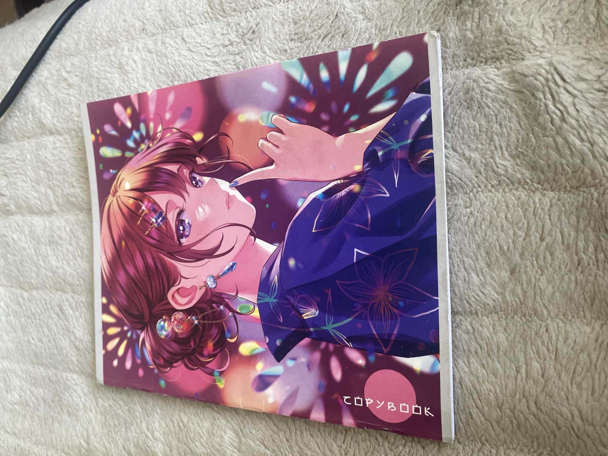 The image shows the cover of a notebook or artbook featuring an anime-style illustration of a young woman with auburn hair styled in an updo, wearing a dark blue yukata with a floral pattern. She has large purple eyes and is looking to her right, with her index finger touching her lips. The background is a vibrant display of colorful, abstract shapes resembling fireworks or bokeh lights. On the left side of the notebook's cover, vertically oriented Japanese text is visible. The notebook is placed on a textured, light-colored surface, possibly a blanket or rug. There are no discernible elements in the image that would suggest a location in Krupki, Belarus.
